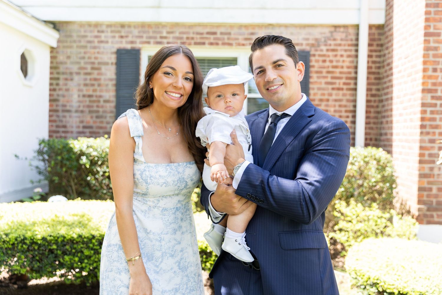 A smiling couple poses outdoors holding their baby dressed in white christening attire near a brick building.