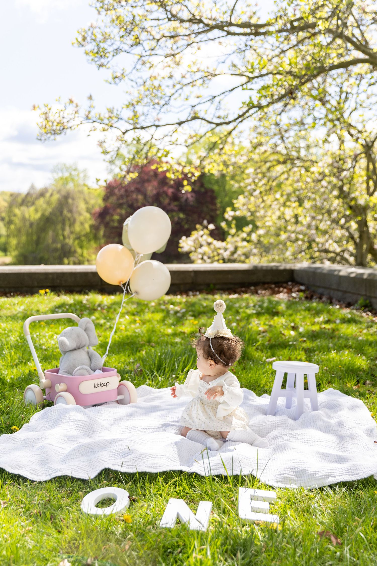 Baby girl in white dress sitting on blanket with ONE letters, balloons, and toy stroller outdoors.