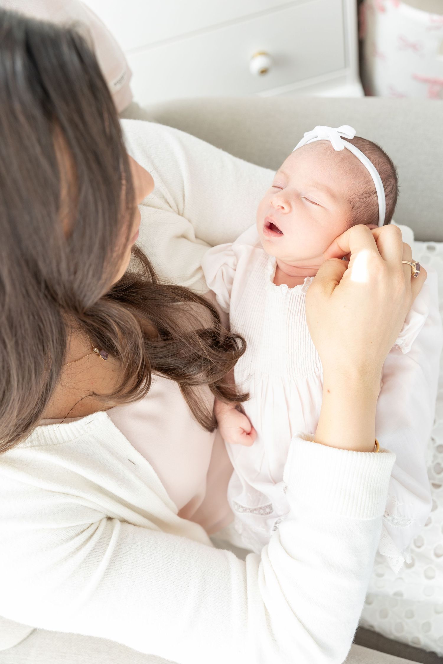 Mother in white cardigan gently touching cheek of crying newborn baby girl with white headband.