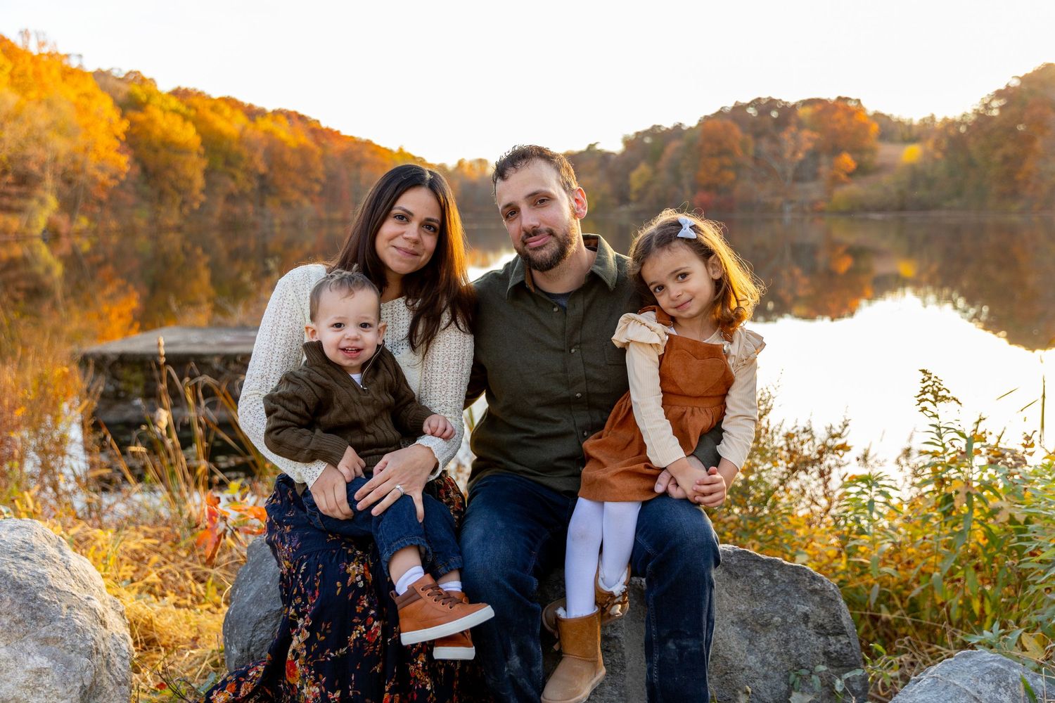 Family of four posing on rocks by a lake surrounded by colorful autumn foliage.