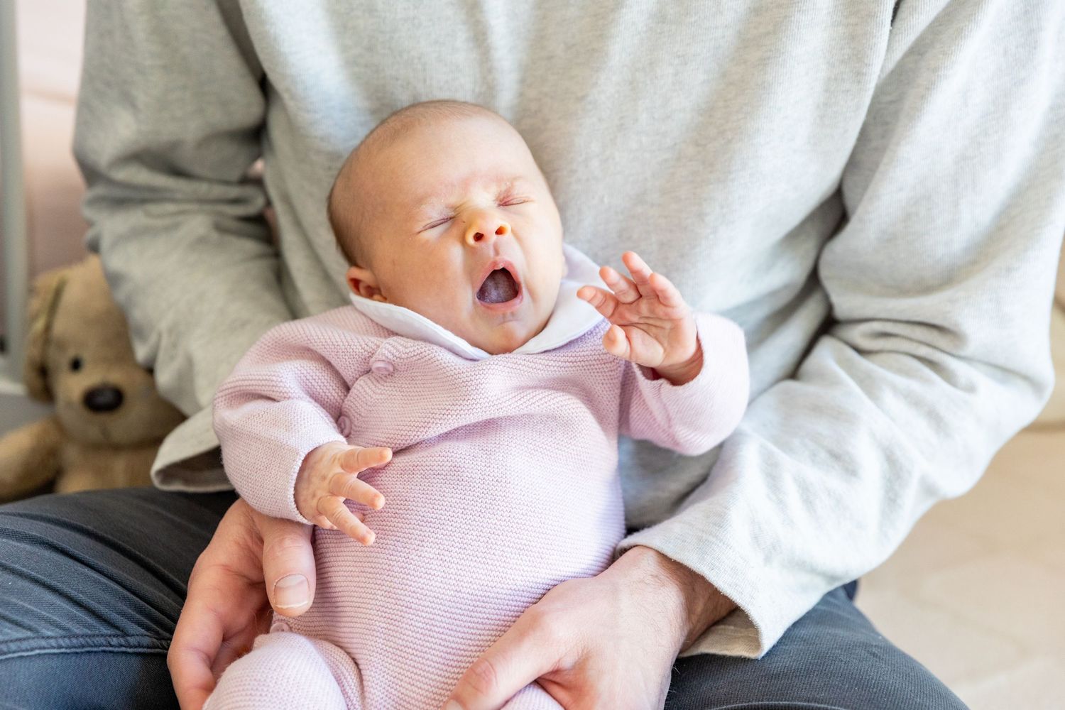 Adorable newborn baby in pink outfit yawning while being held in parent's arms.