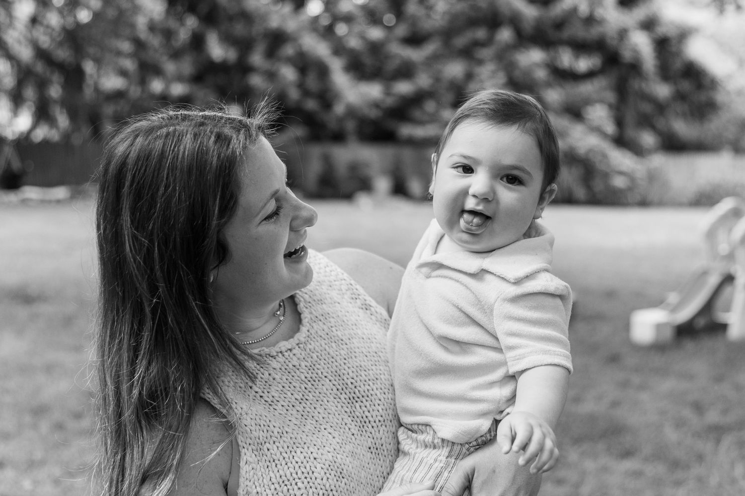 Black and white photo of smiling mother holding happy laughing baby outdoors in garden setting.