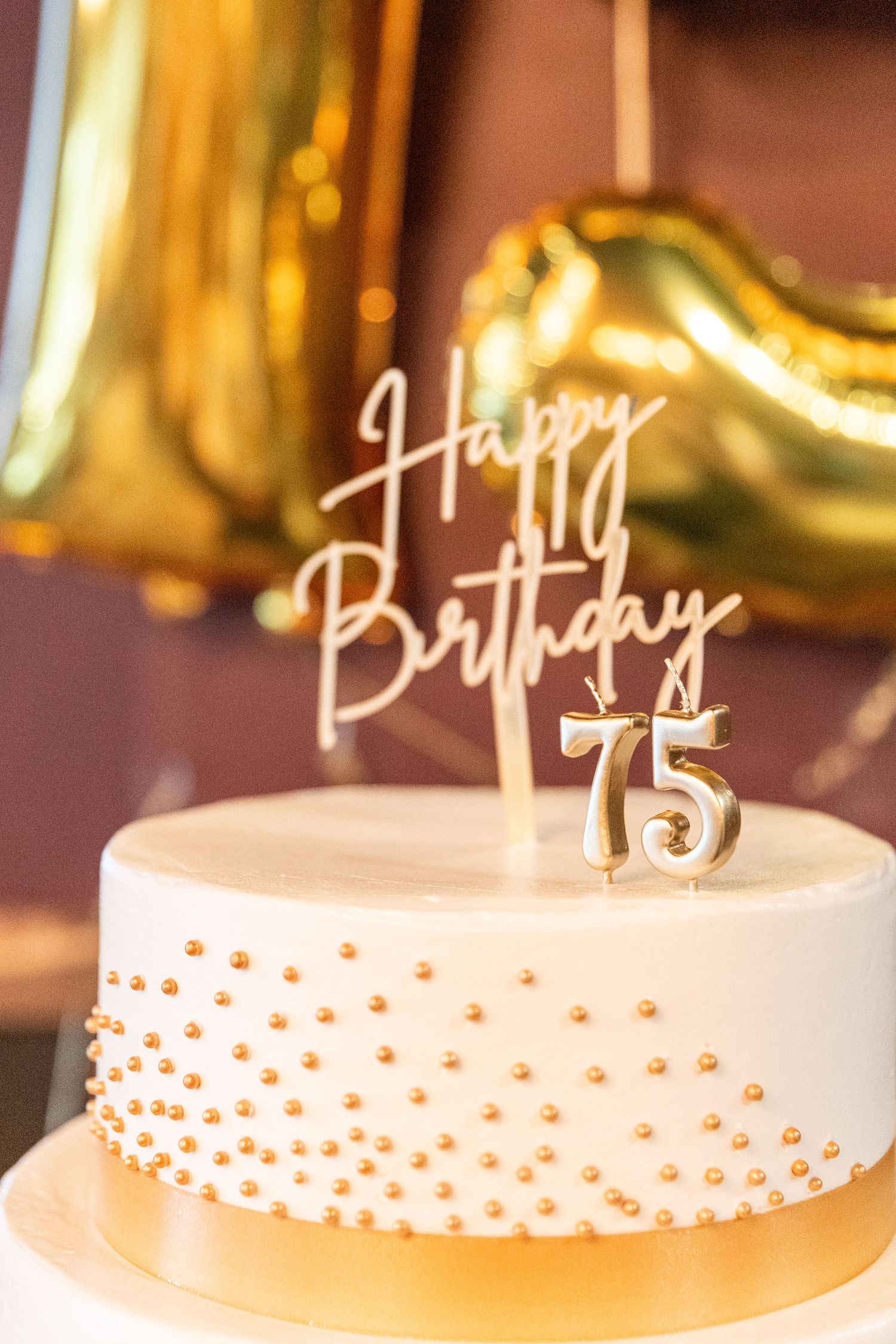 Elegant white birthday cake with gold pearl details and Happy Birthday 75 topper with gold balloons.