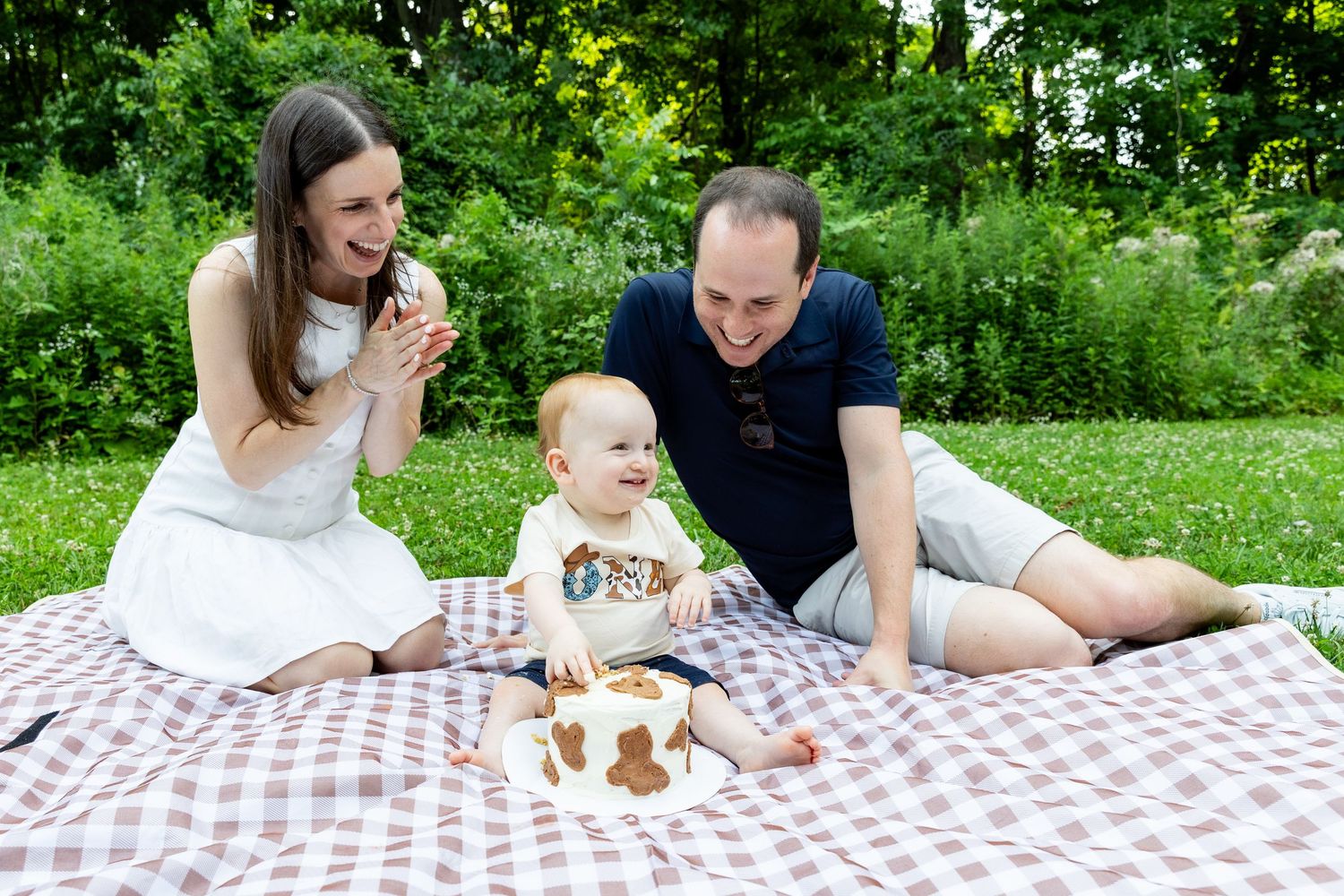 Happy family celebrating baby's first birthday with cowboy cake on picnic blanket outdoors.