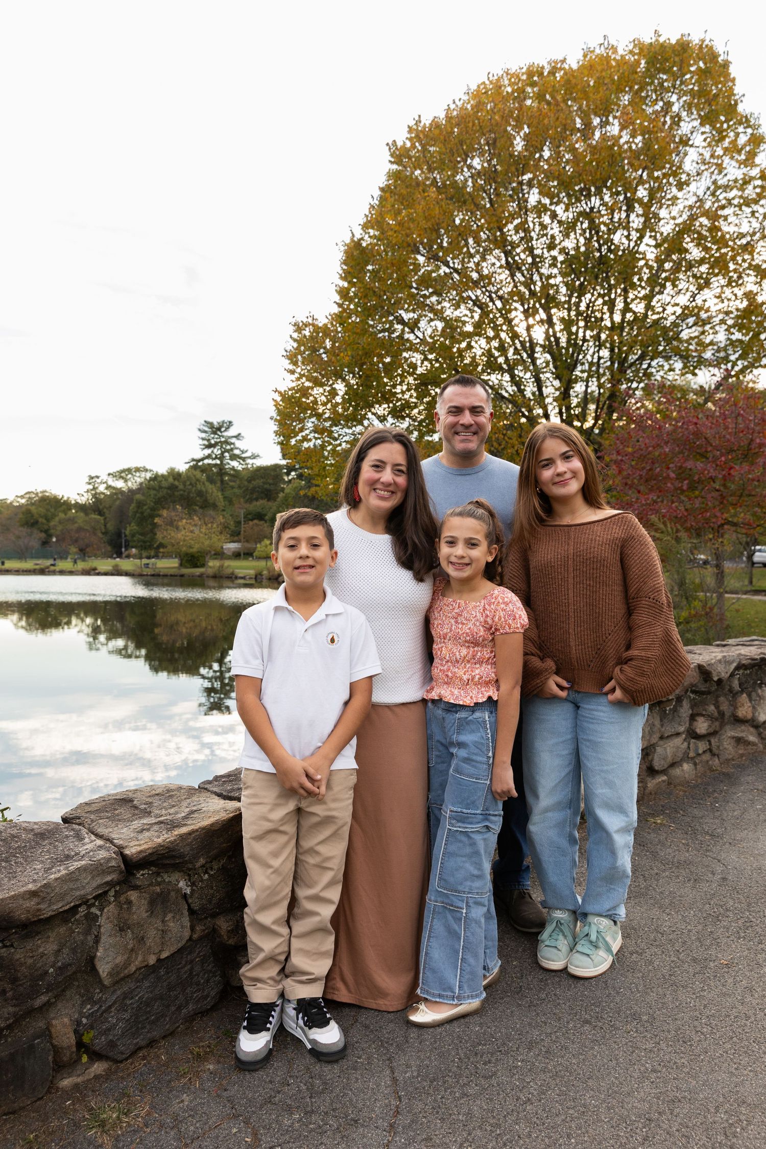 Family of five posing by a pond with autumn trees in a park setting.