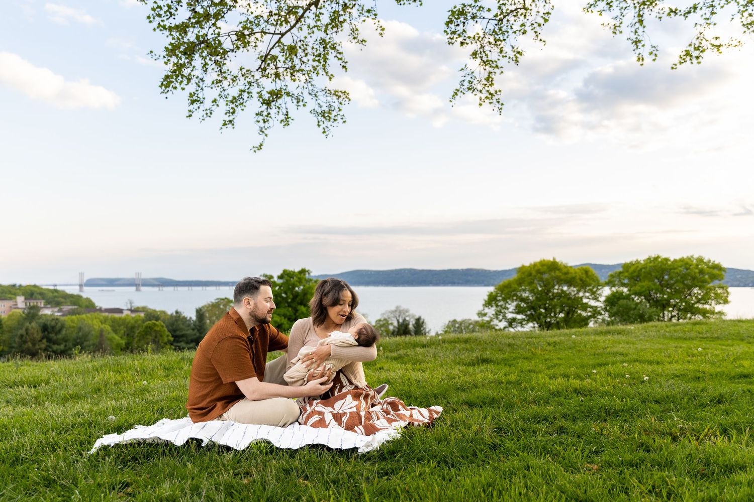 Couple sitting on a blanket on a green hillside cradling a newborn baby at sunset.