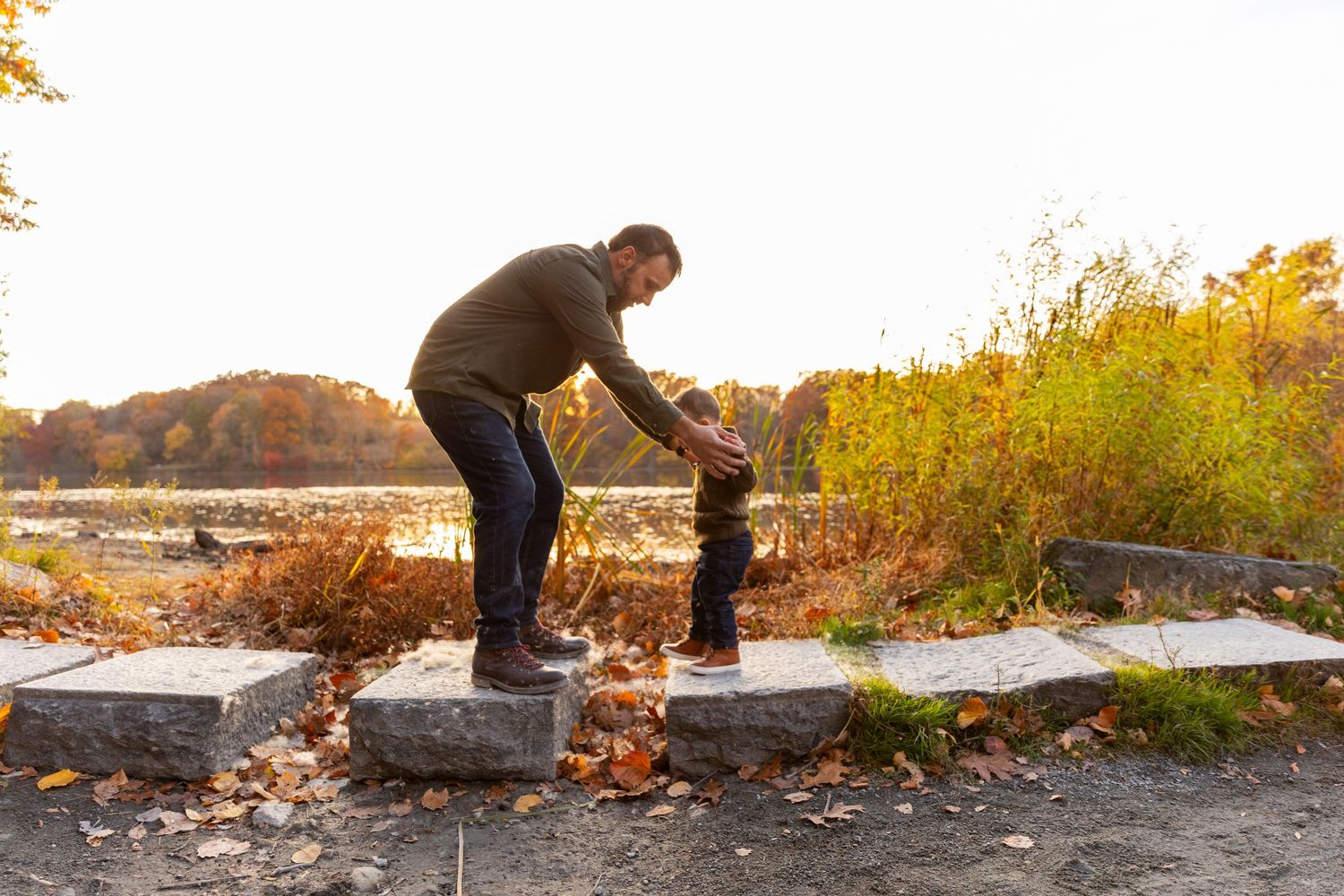 Father helping young toddler walk on stone steps by a scenic autumn lakeside.
