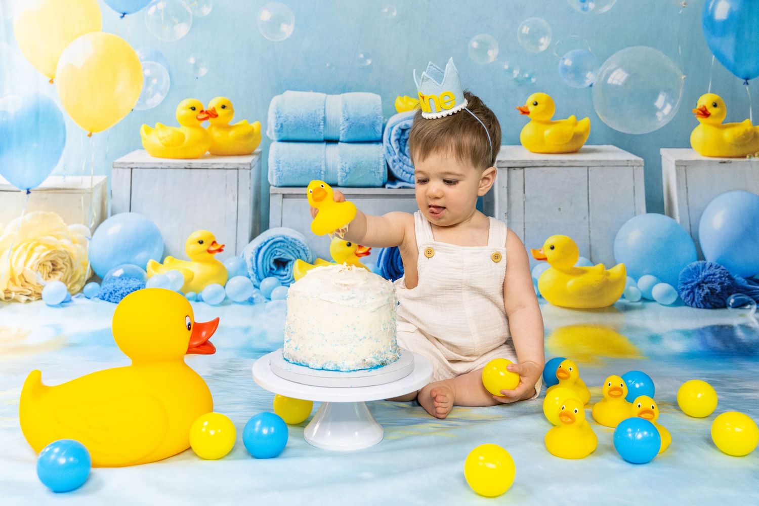 Baby's first birthday cake smash with rubber duck theme, yellow ducks, and blue balloons.