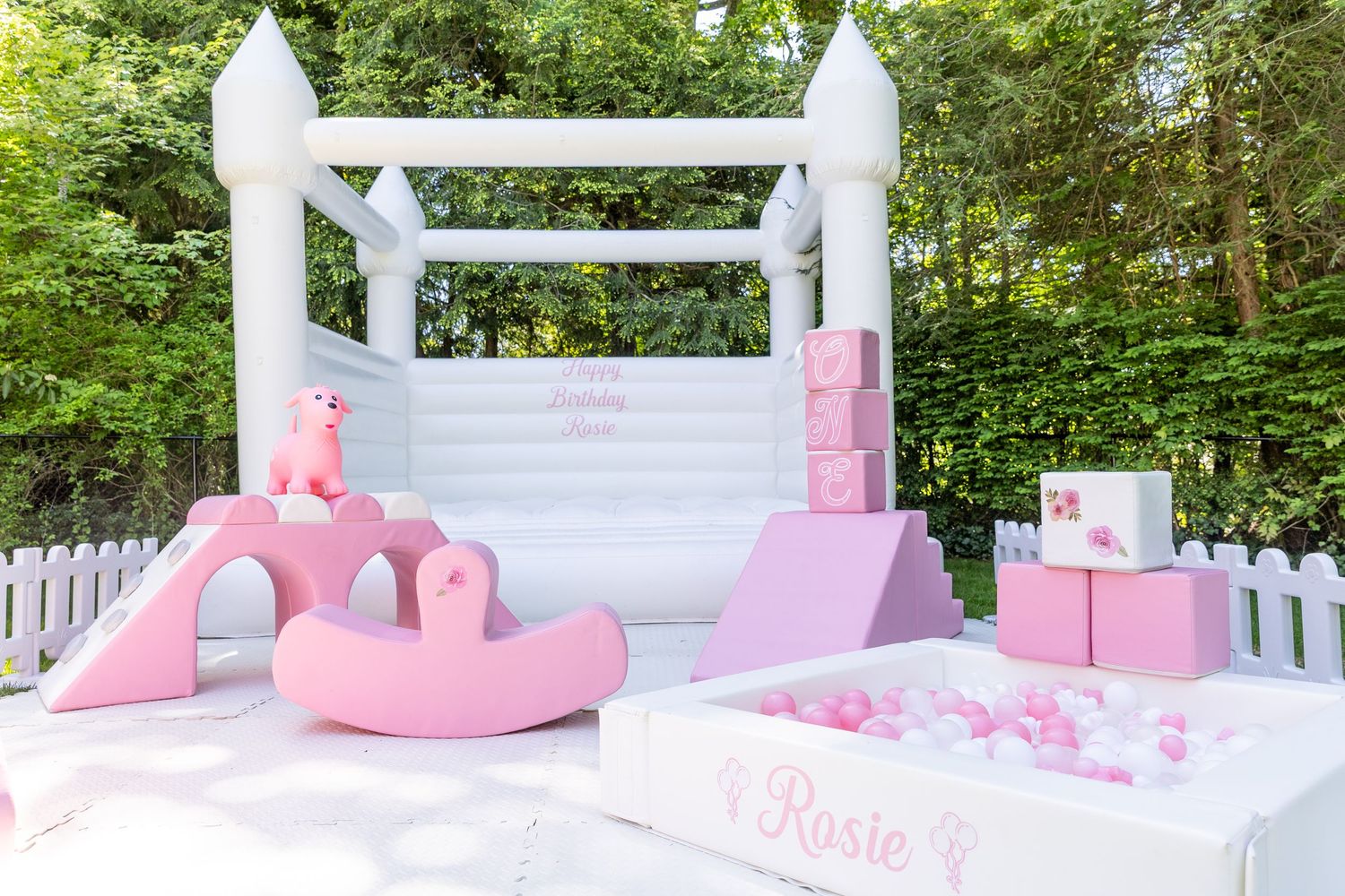 White bounce castle with pink slide and ball pit for a birthday party outdoors.
