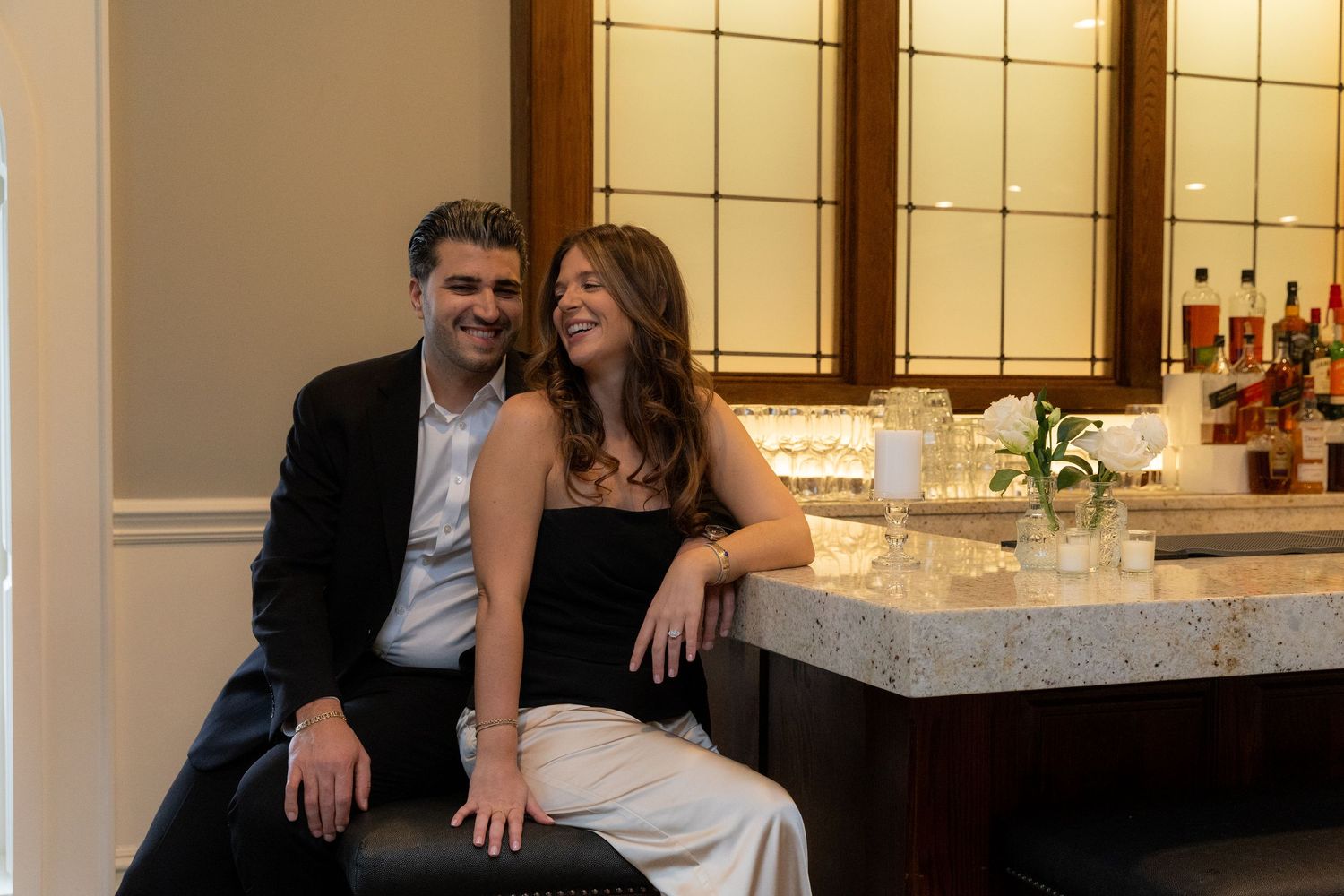 A happy couple sits together smiling at a bar counter in an elegantly lit interior space.