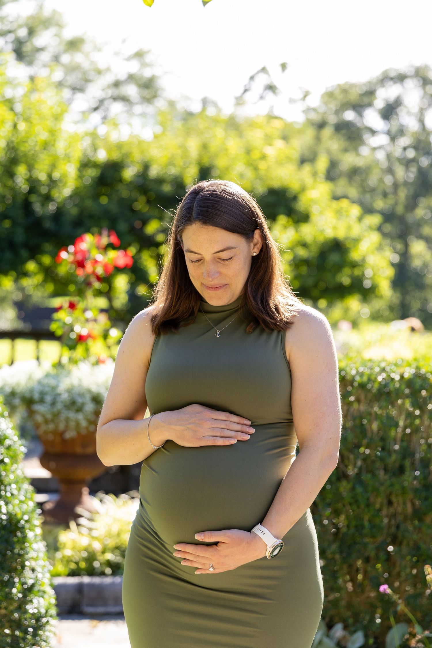 Pregnant woman in olive green dress cradling baby bump in sunny garden with flowers.