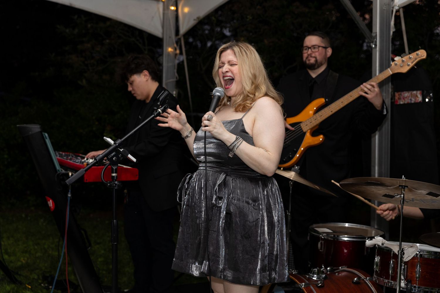 Live band performing under a tent at night with female singer in silver dress holding microphone.