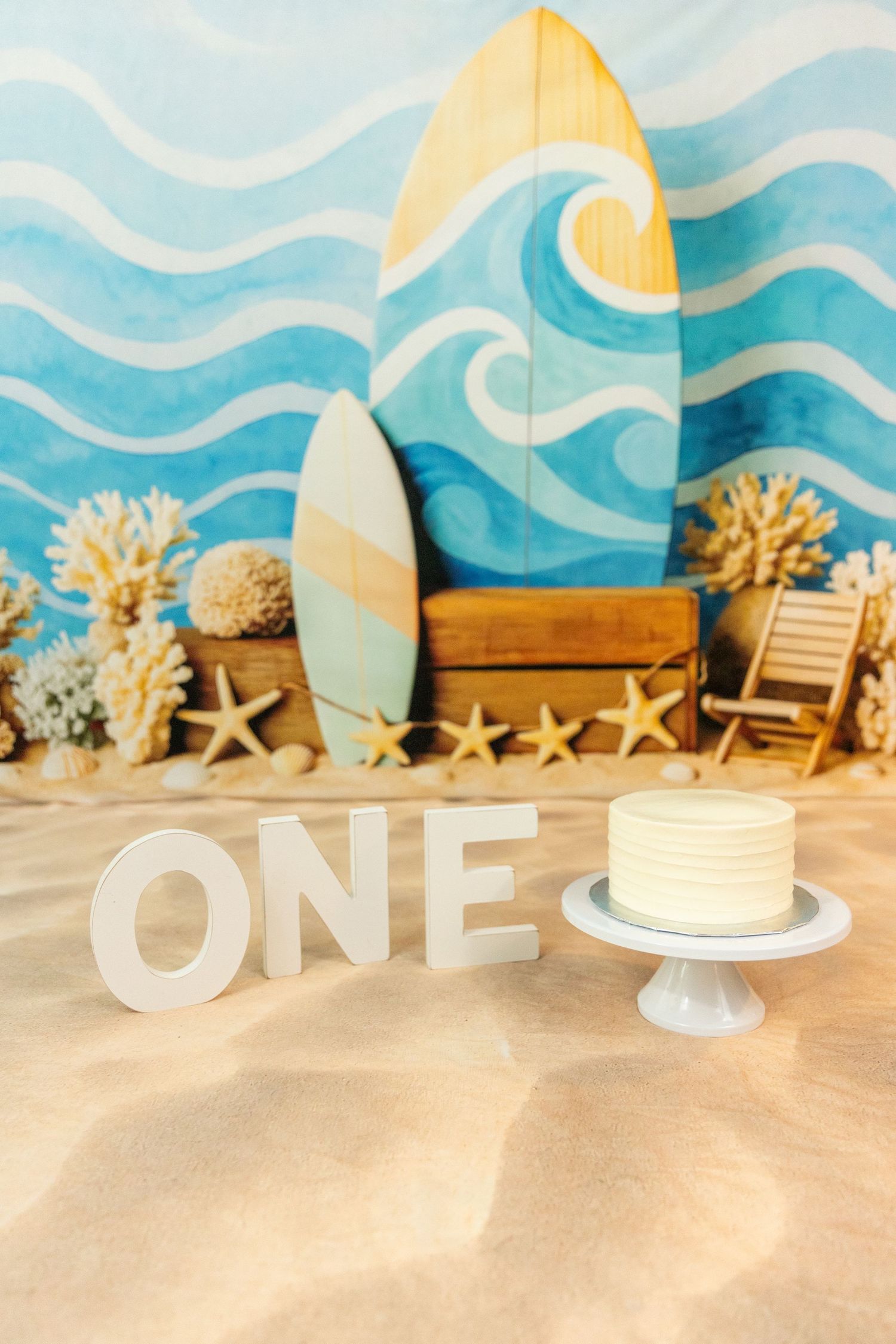 Beach birthday cake smash setup with ONE letters, white cake, surfboards, and ocean wave backdrop.