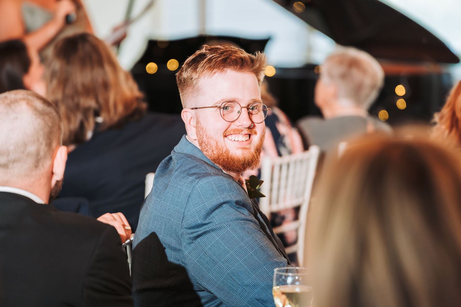 A person with red hair and glasses smiles while sitting at a social gathering with others in the background.
