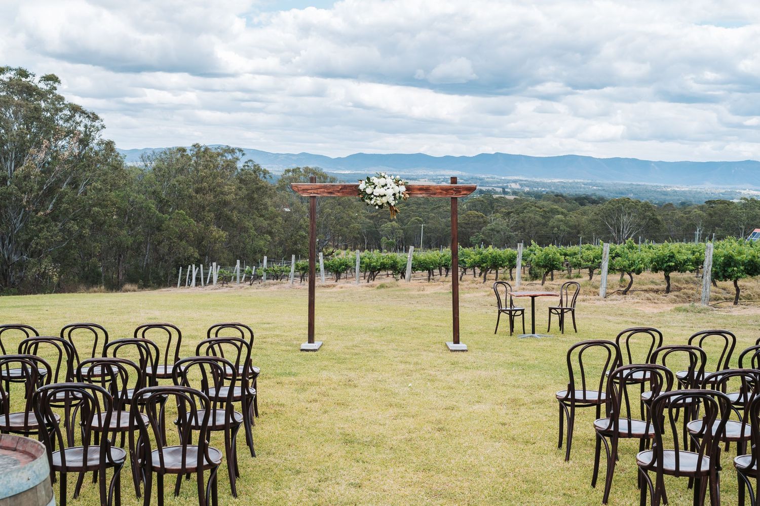 Outdoor wedding ceremony setup with black chairs facing a wooden arch overlooking a vineyard landscape.