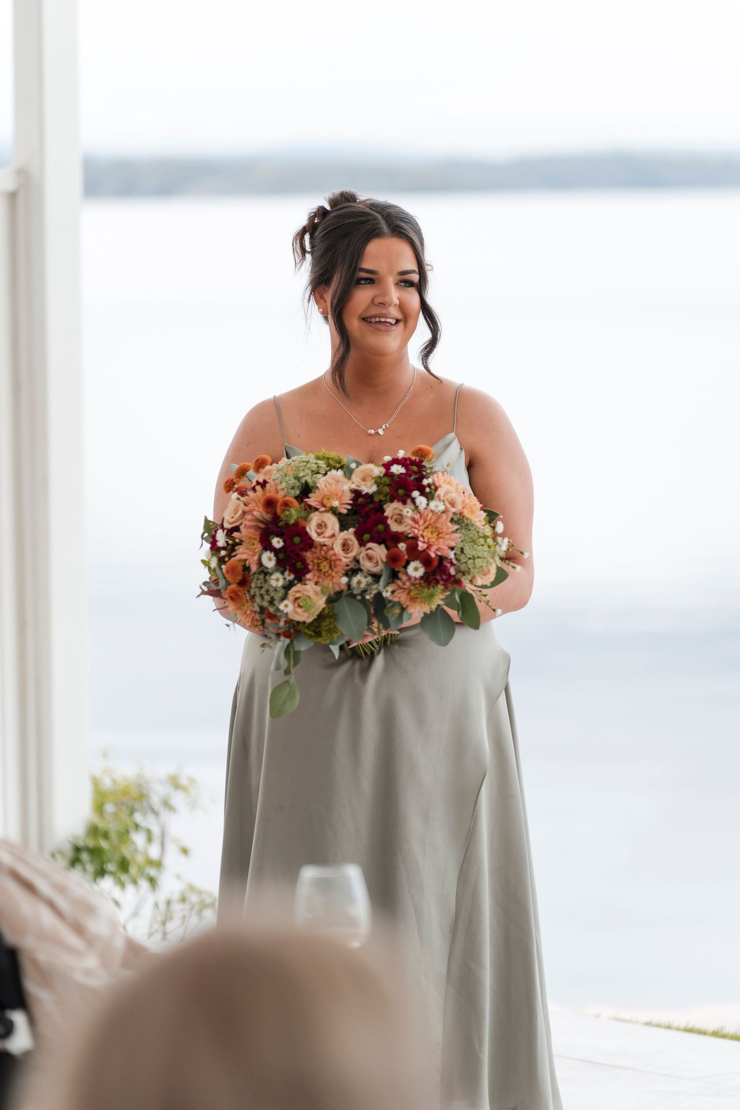 A bridesmaid in silver dress holds a lush coral and peach bouquet against waterfront backdrop.