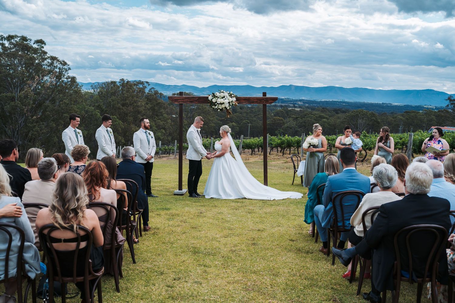 Outdoor wedding ceremony on hilltop with mountain views and guests seated on white chairs.