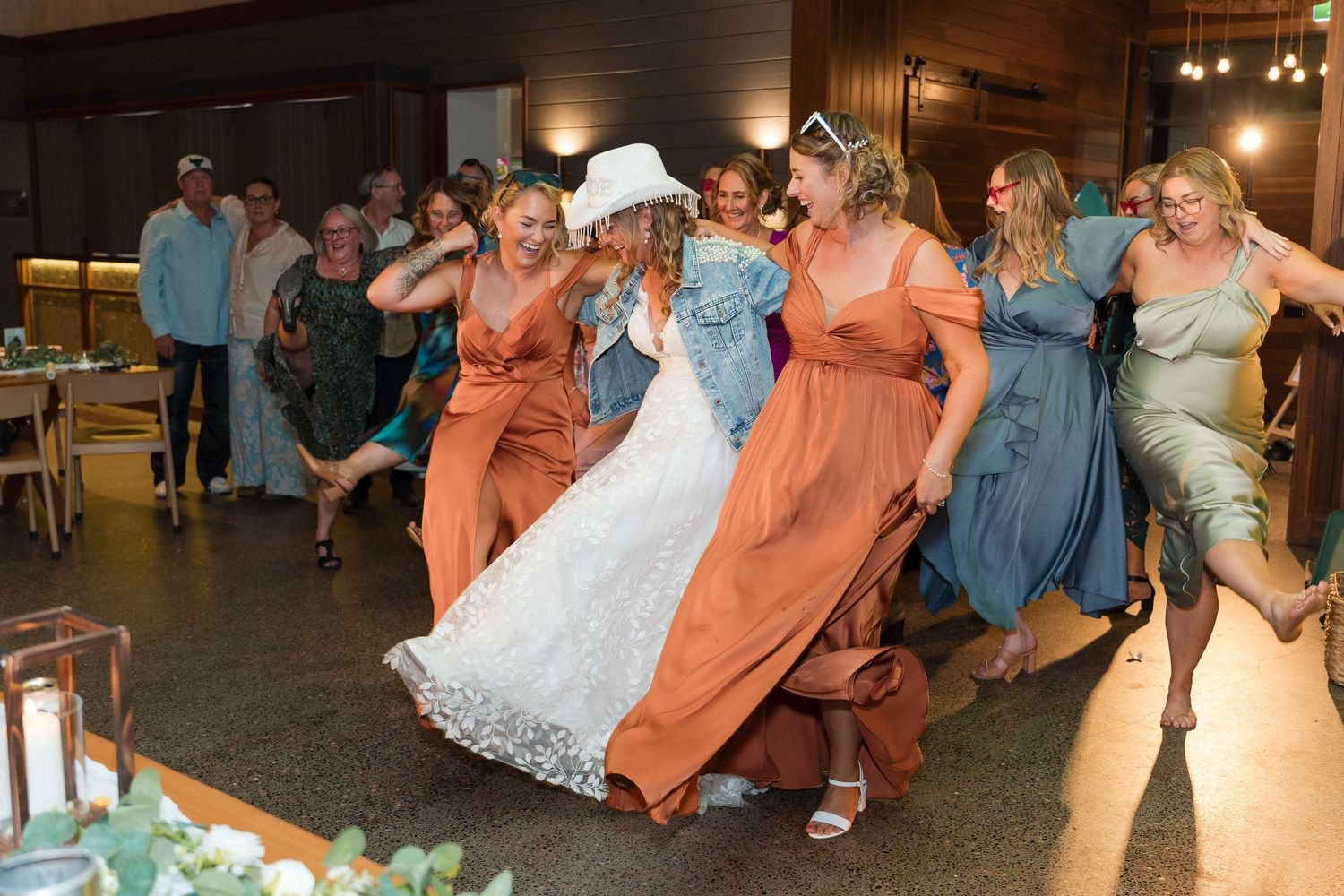 Bride in white lace gown and cowboy hat dances energetically with bridesmaids in orange and blue dresses.
