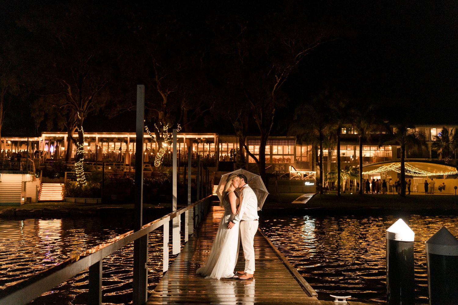 Newlyweds share romantic kiss under umbrella on waterfront dock at night with illuminated venue behind them.