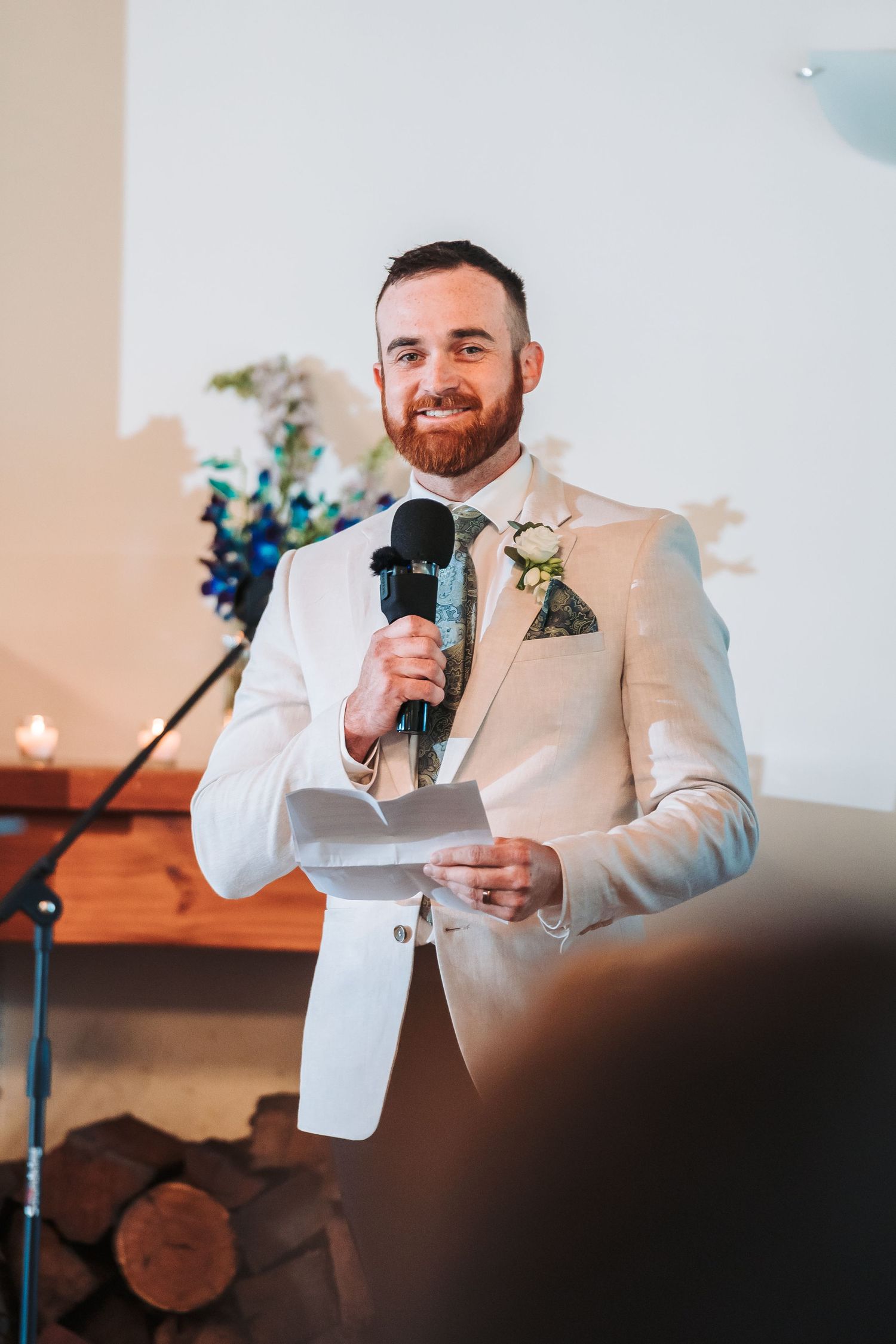 A groom in a white suit gives a wedding speech while holding a microphone and notes during an indoor reception.