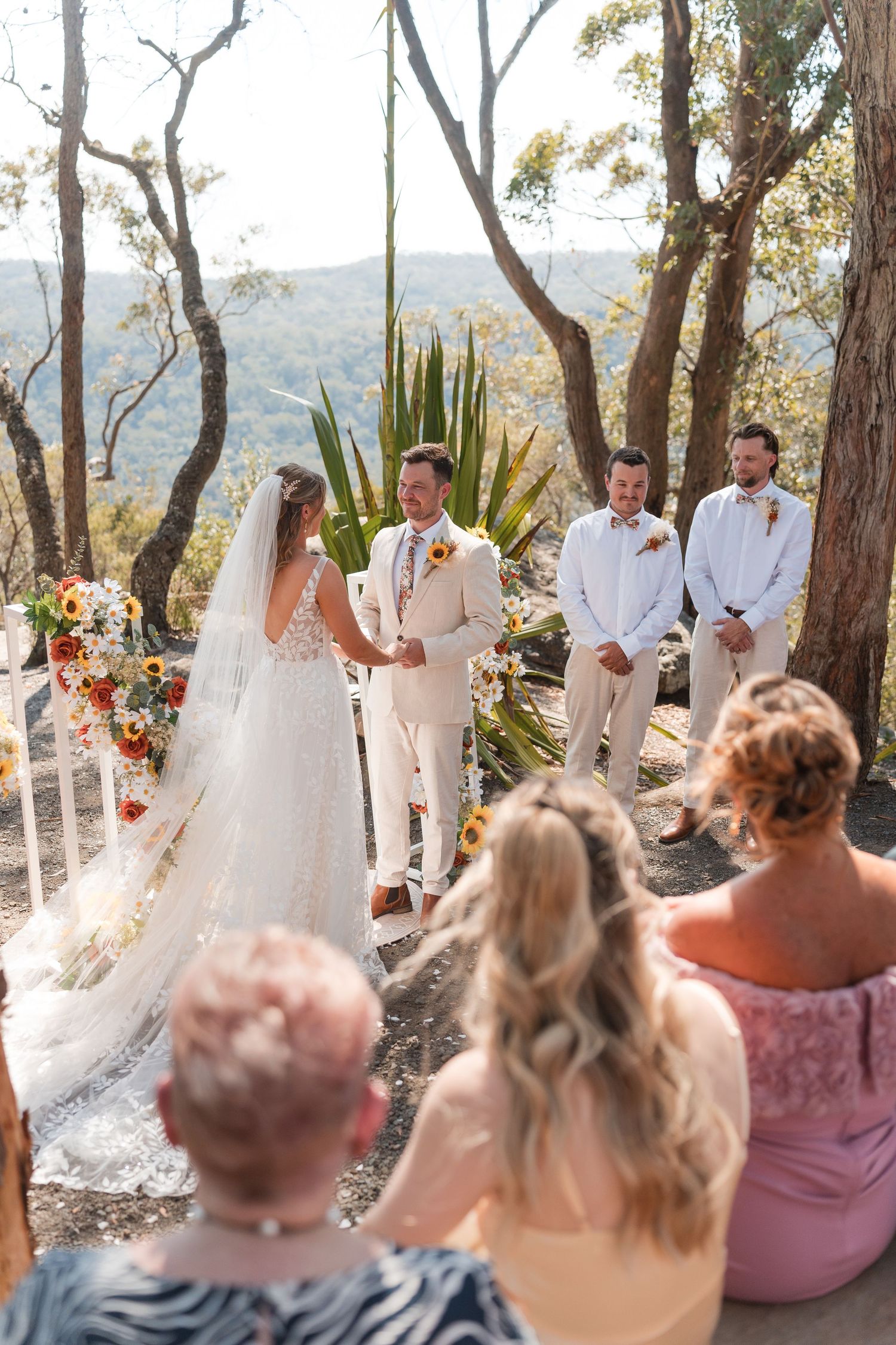 Outdoor wedding ceremony in bushland with bride and groom holding hands at altar, groomsmen standing nearby, guests seated.
