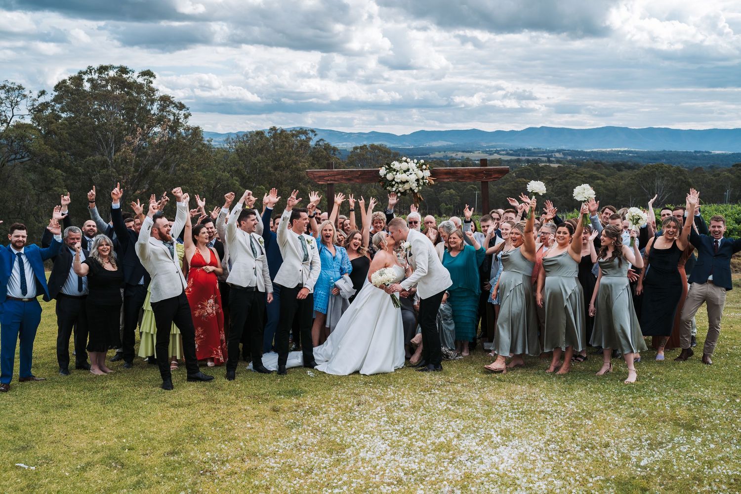 Large wedding group poses for celebration photo on hilltop with mountain views.