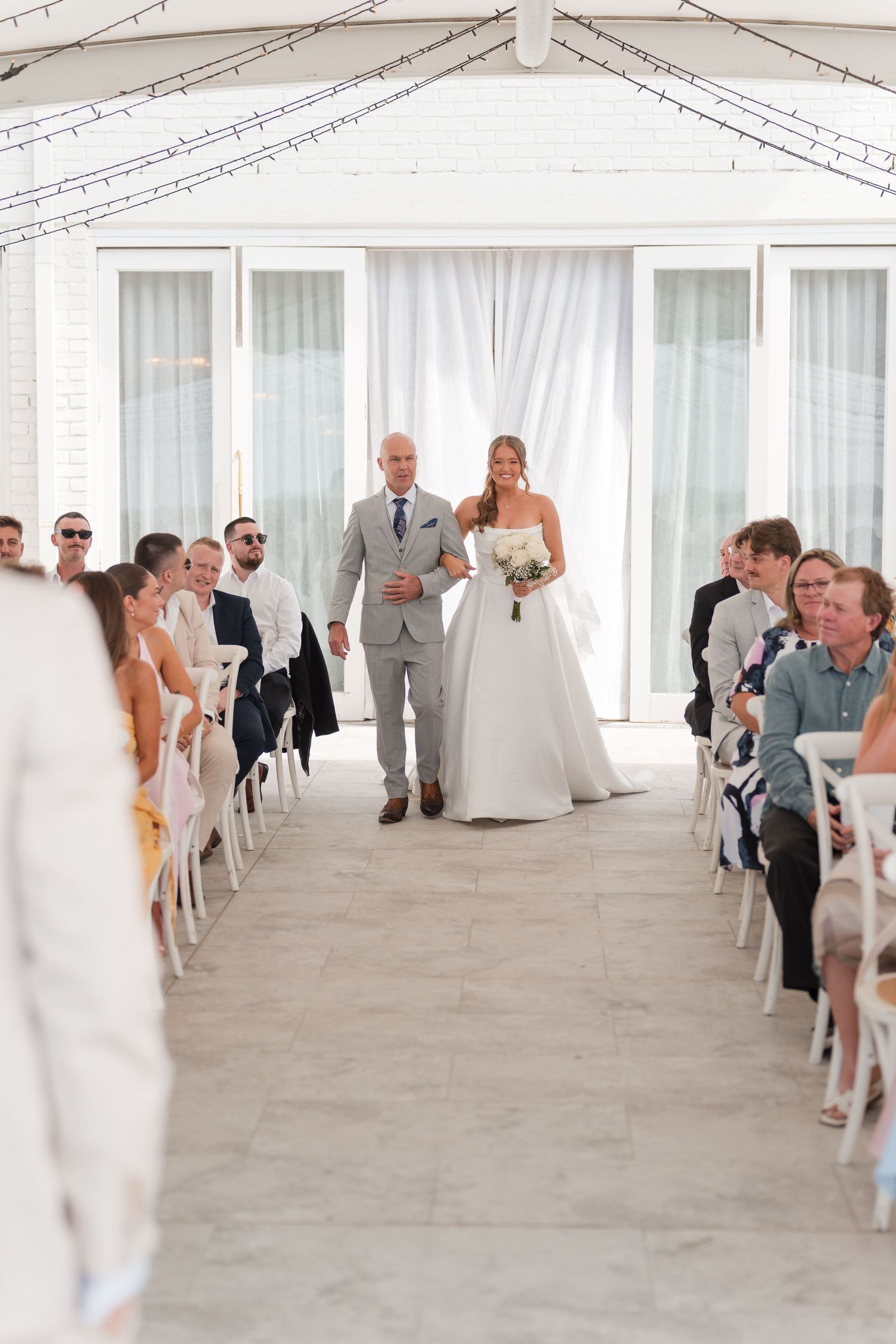 Bride in white ball gown walks down aisle with escort at elegant indoor wedding ceremony.