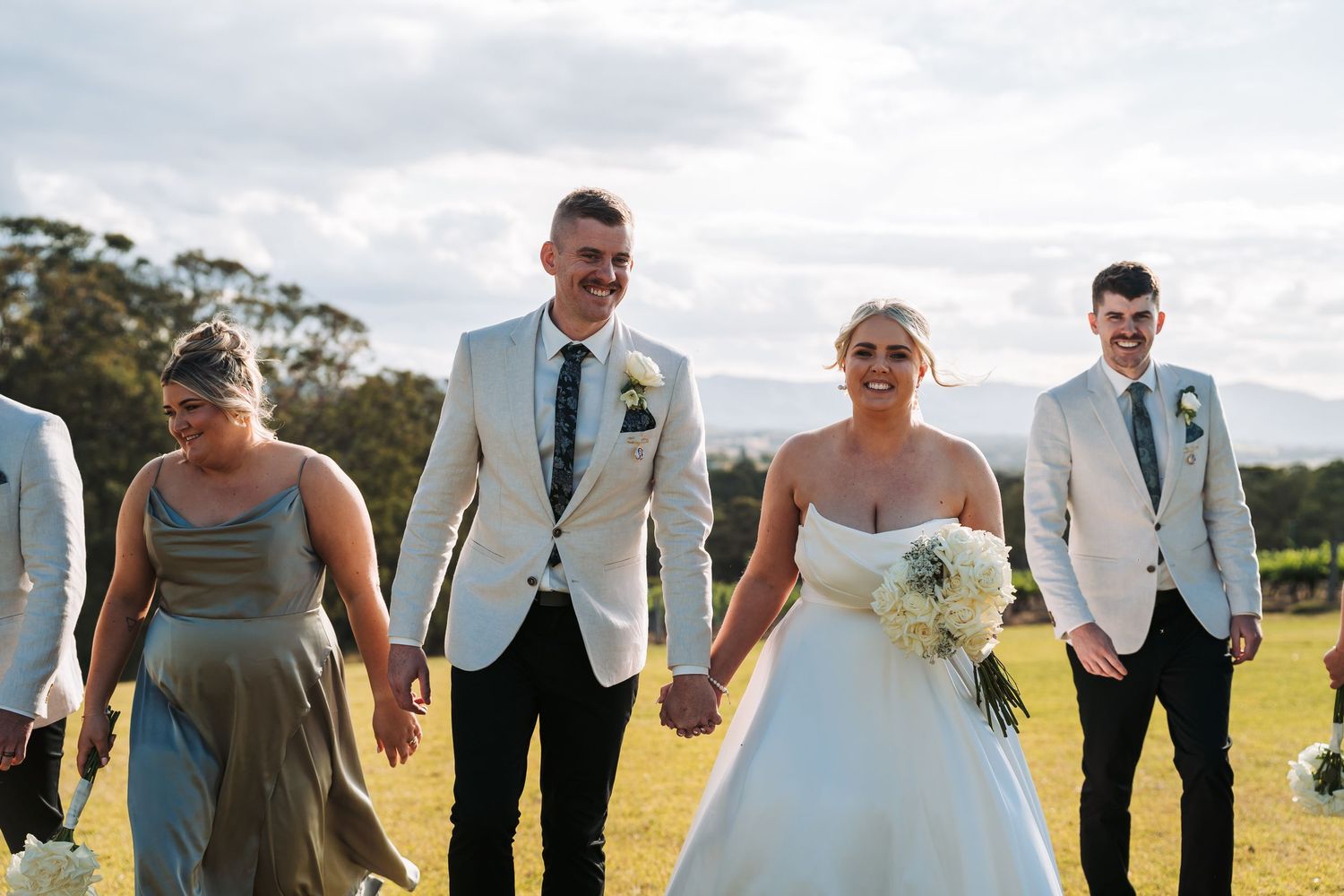 Wedding party walks together across a grassy field with a scenic vineyard backdrop.