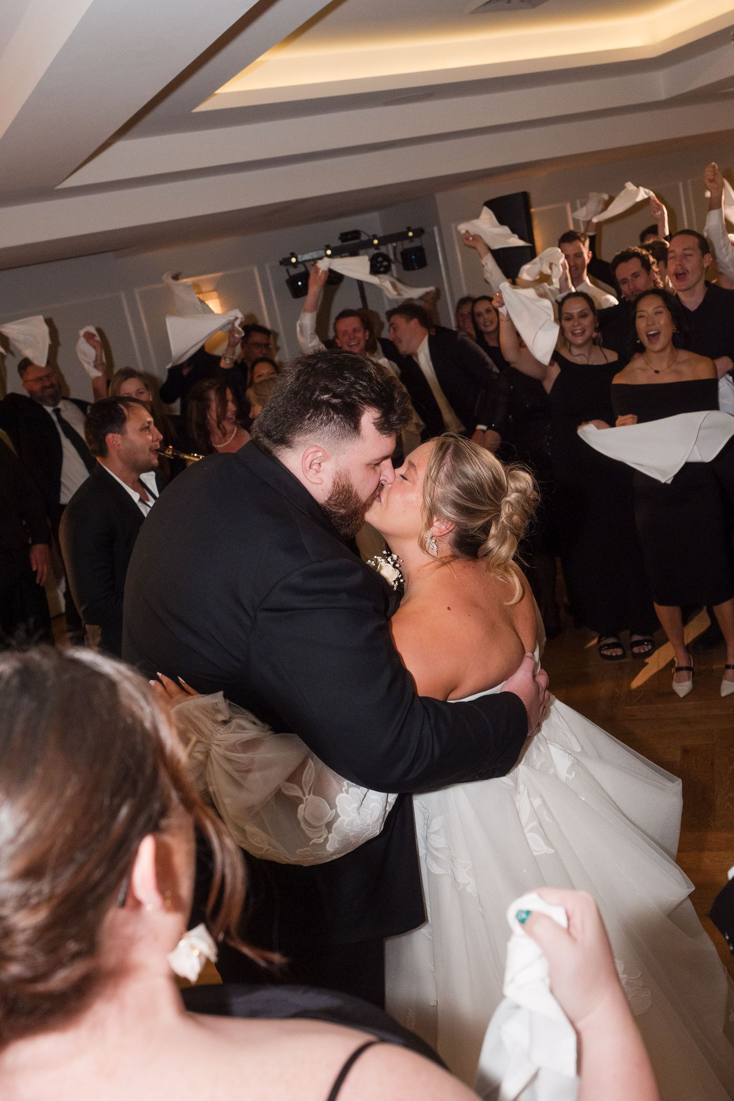 Bride and groom share a passionate kiss on the dance floor surrounded by cheering guests waving napkins at wedding reception.