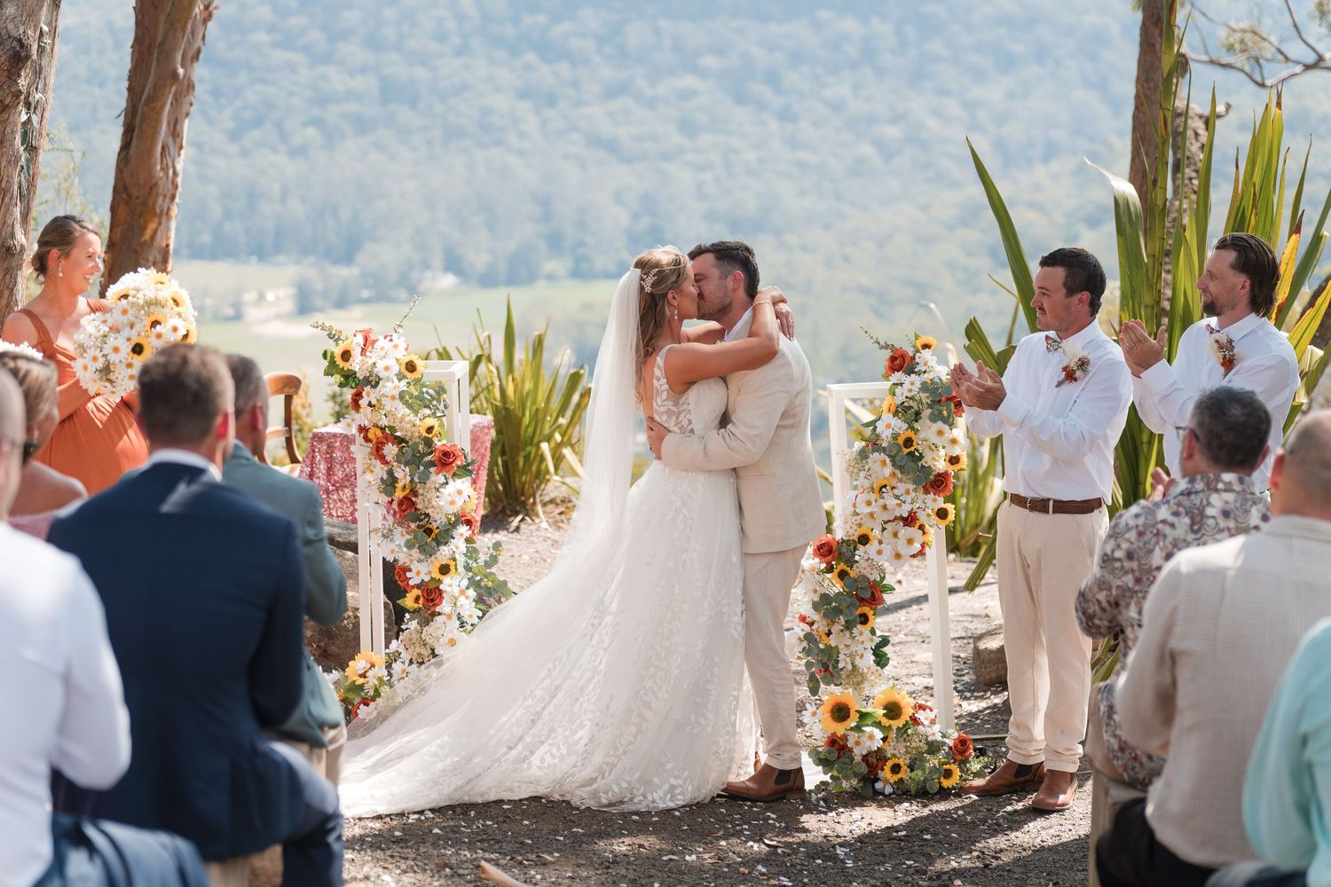 A bride and groom share their first kiss at an outdoor wedding ceremony surrounded by sunflower arrangements.