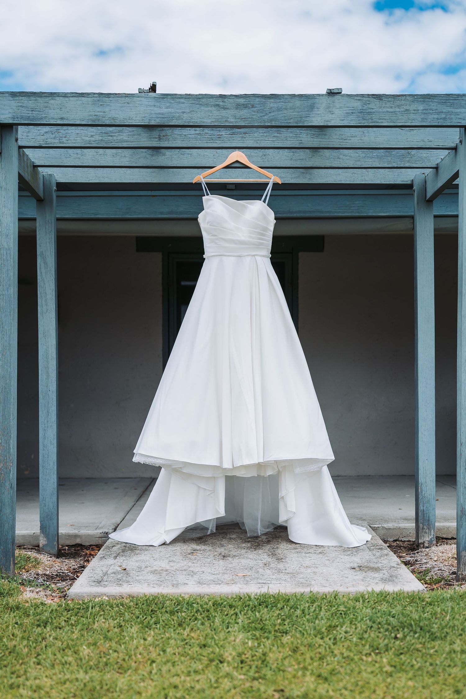 A white wedding dress hanging on a wooden hanger under a blue-gray pergola against a cloudy sky.