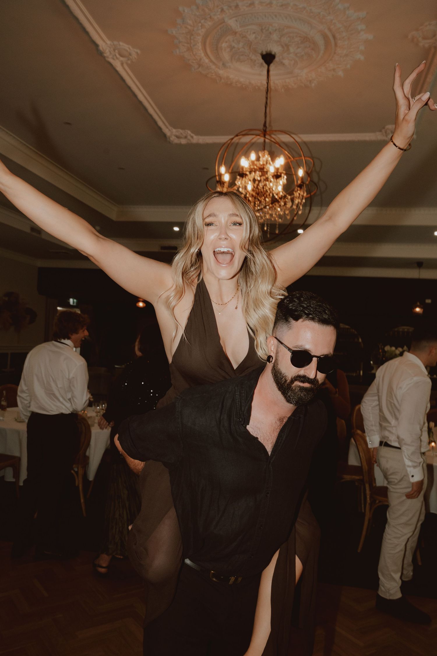 Excited blonde woman with arms raised sitting on bearded mans shoulders at wedding reception.