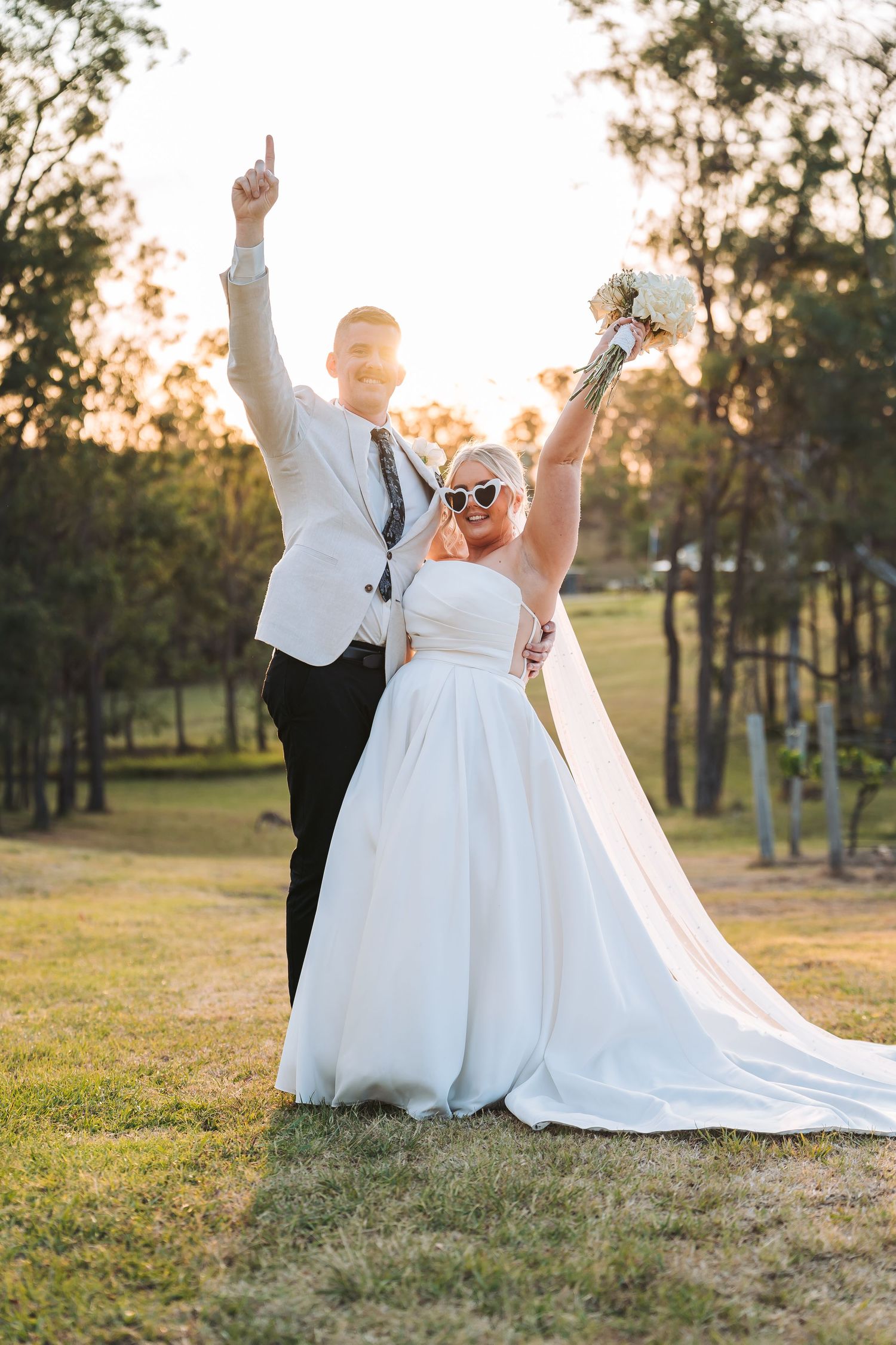 A joyful wedding couple celebrates during golden hour outdoors with raised arms and beautiful sunset backlighting.