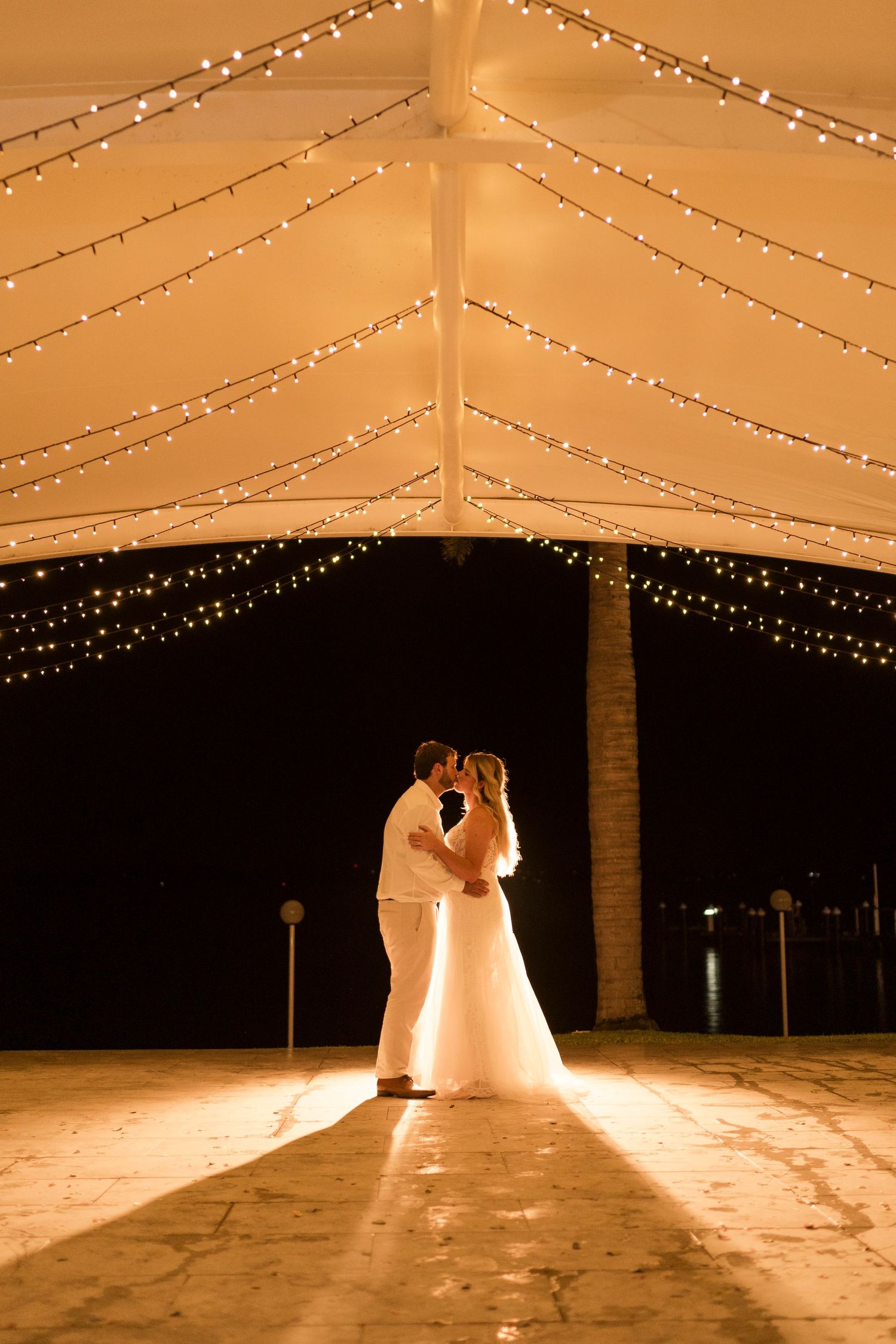 A dramatic backlit silhouette of newlyweds sharing a tender moment under string lights in a wedding tent.
