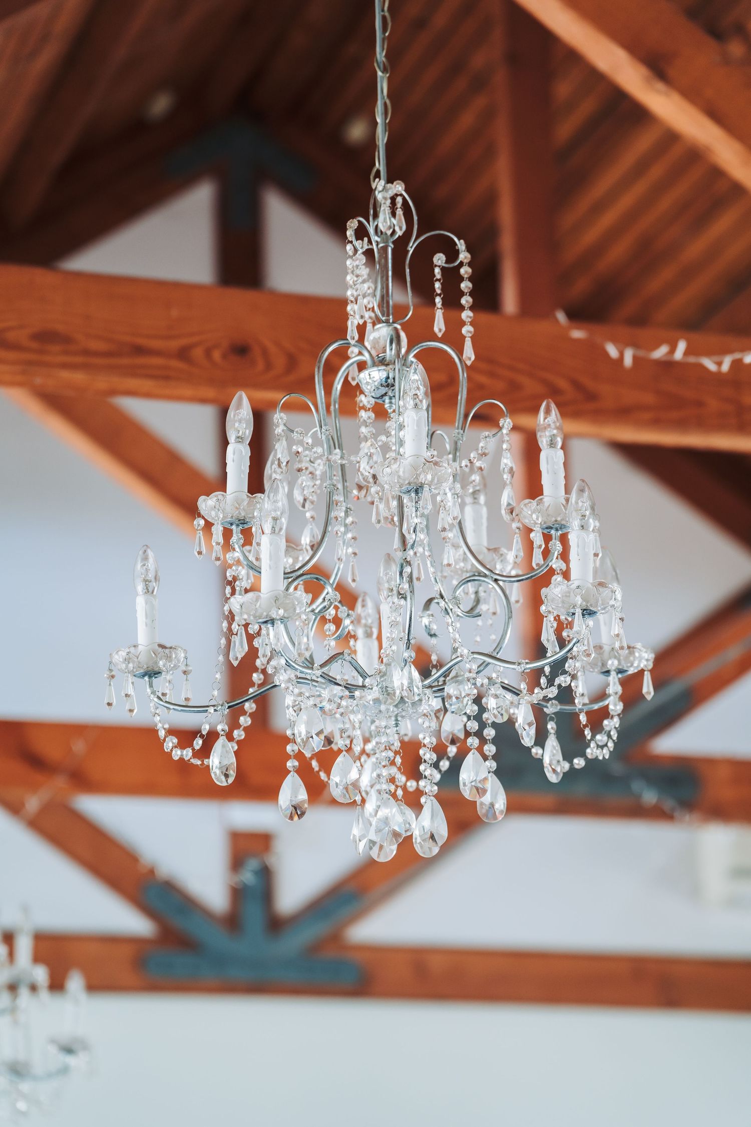 An elegant crystal chandelier with multiple tiers hangs from exposed wooden beams in an airy white room.