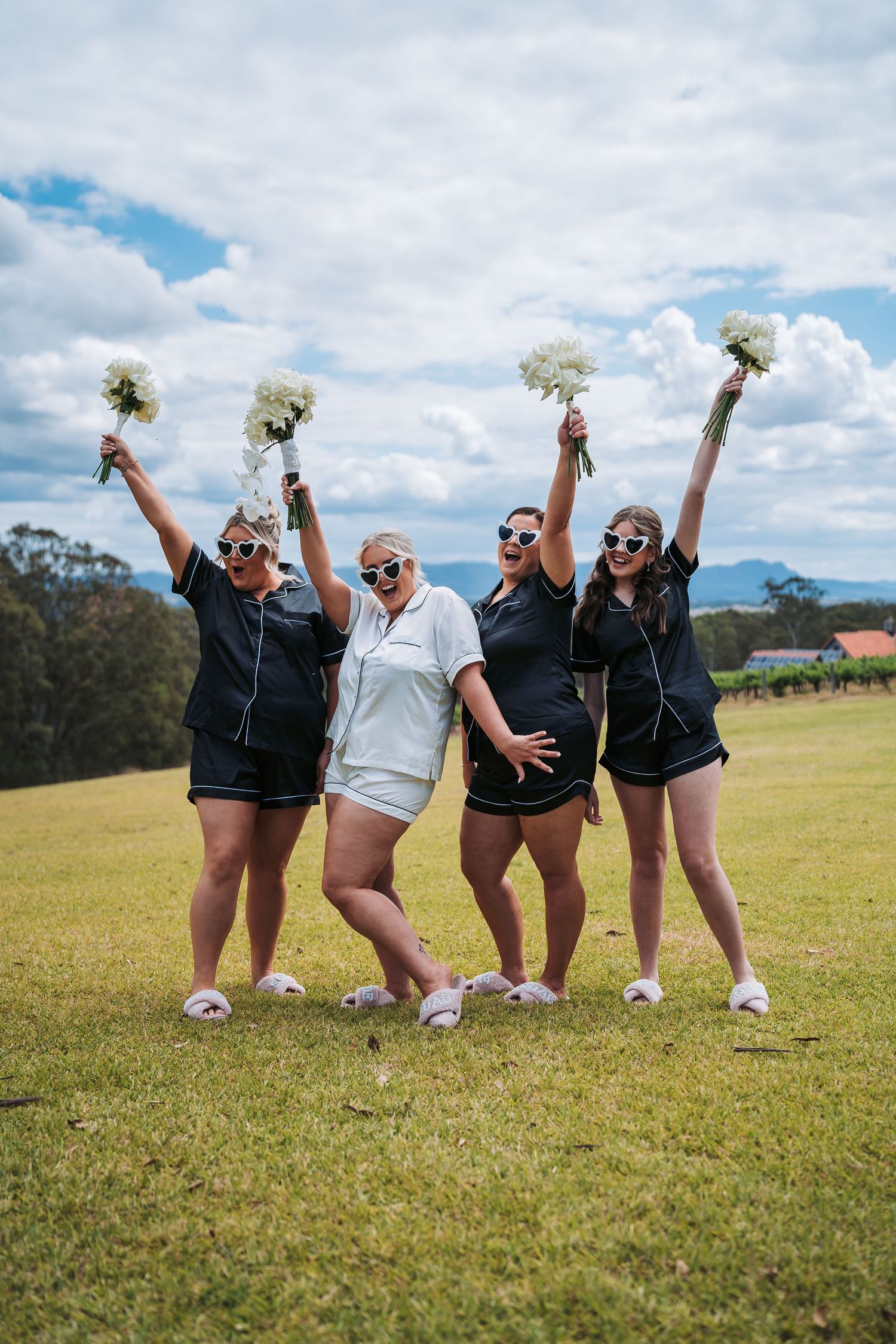 Group of bridesmaids in matching outfits celebrating with bouquets against cloudy sky.