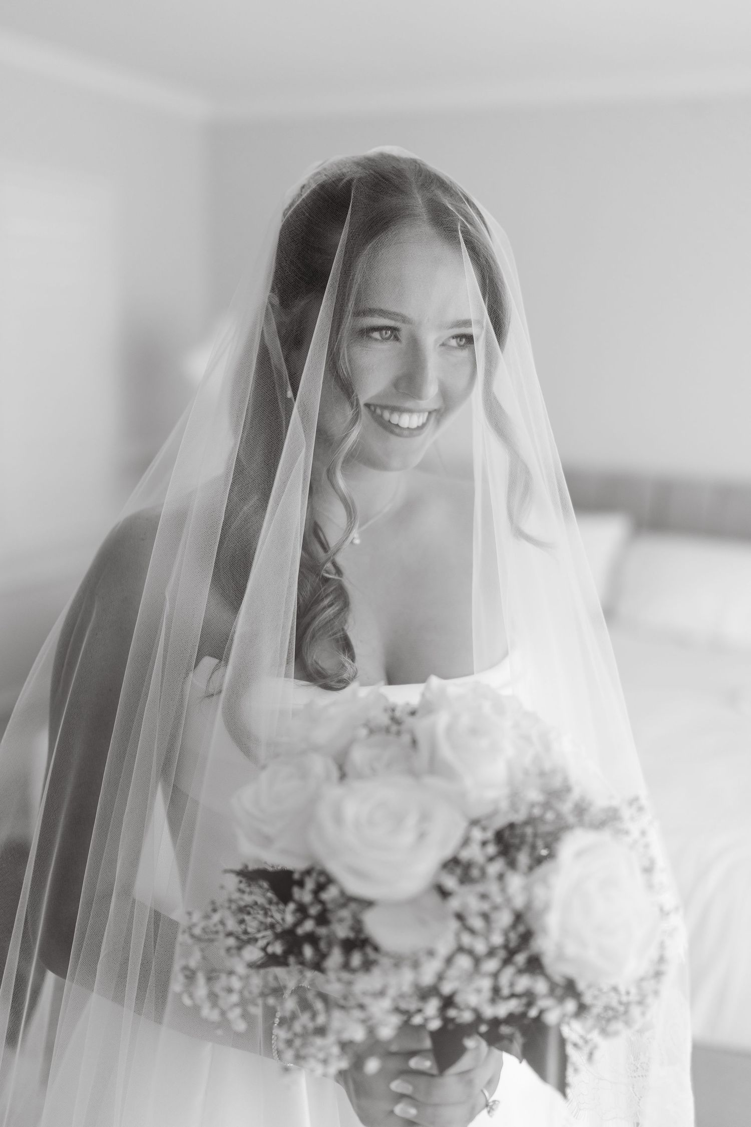 Smiling bride wearing a veil holding a white rose bouquet in a black and white wedding portrait.