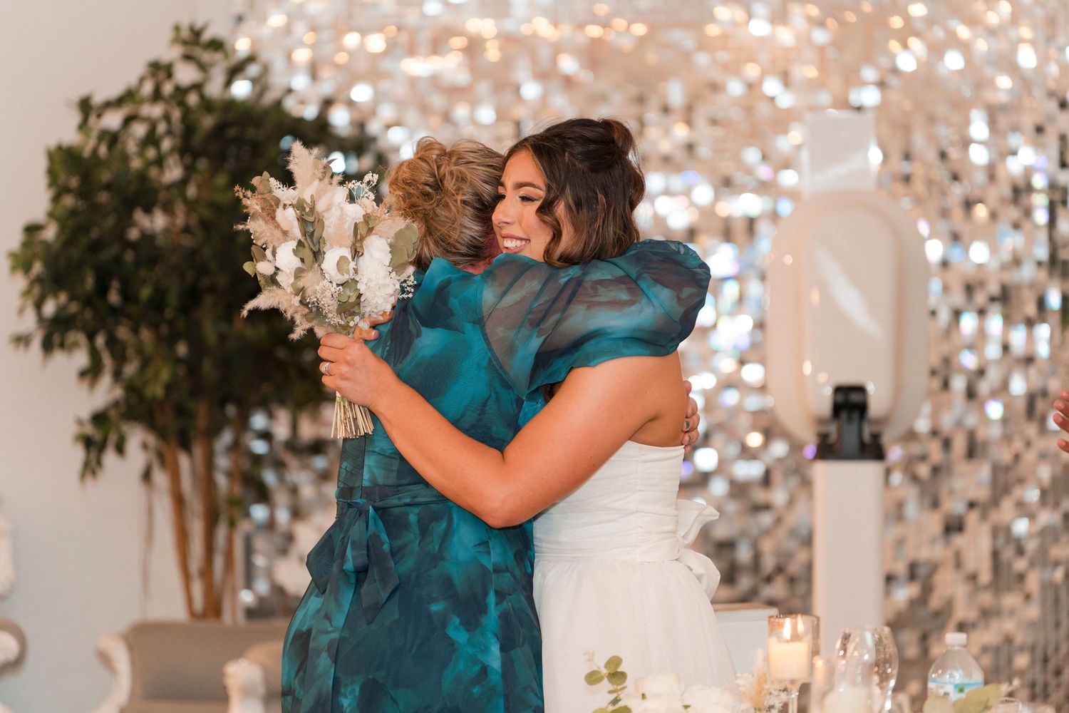 Two people in formal attire share an emotional embrace during a celebration with sparkly backdrop and greenery decor.