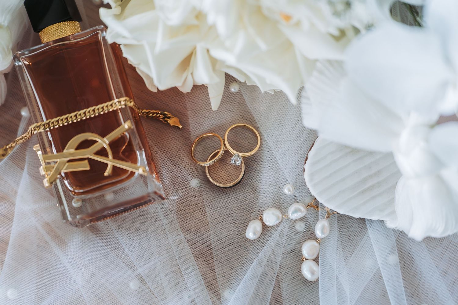 Wedding details featuring gold rings, perfume bottle, and pearls on white fabric.