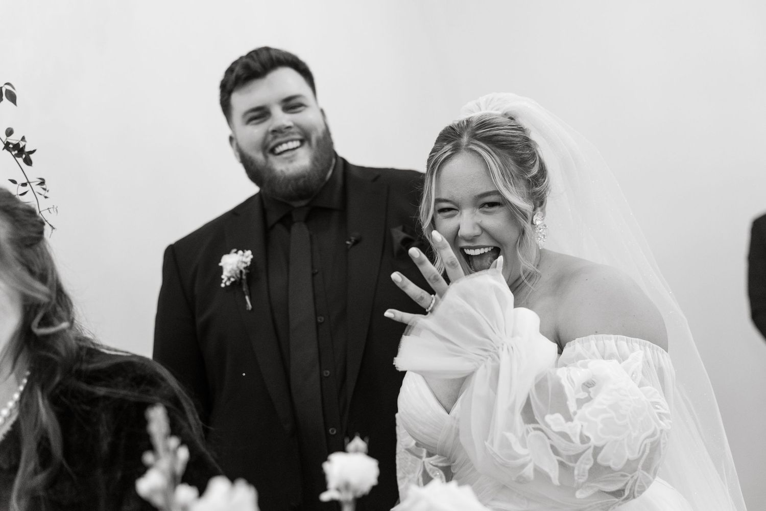 Bride laughs joyfully showing her ring while groom smiles in black tuxedo at wedding ceremony.