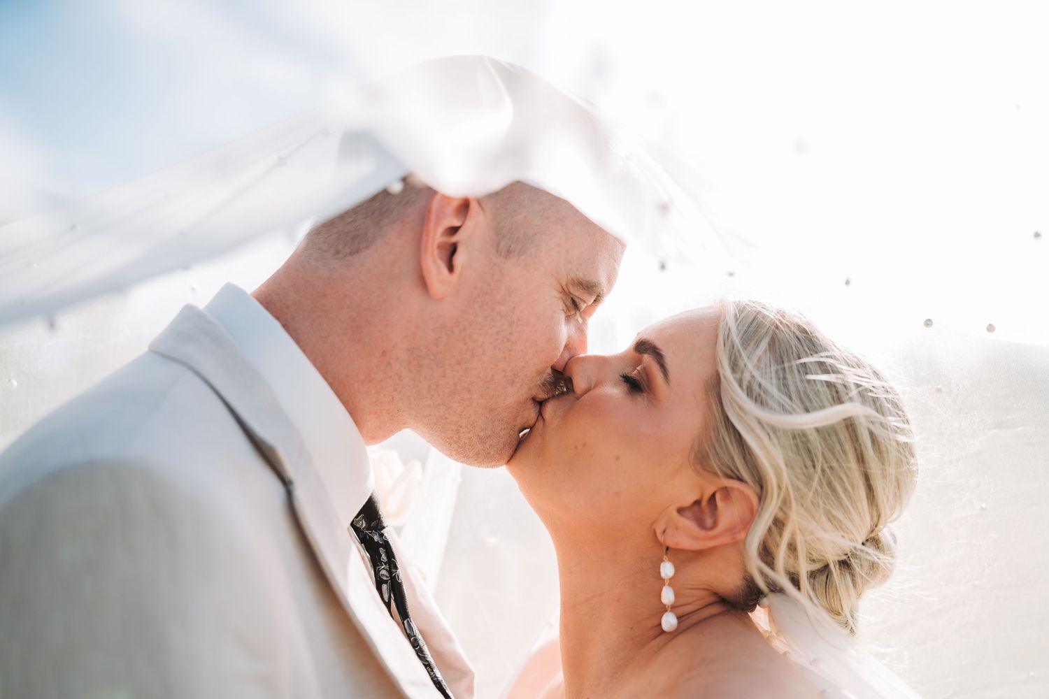 A romantic close-up kiss between newlyweds outdoors with sunlight creating a soft dreamy glow effect.