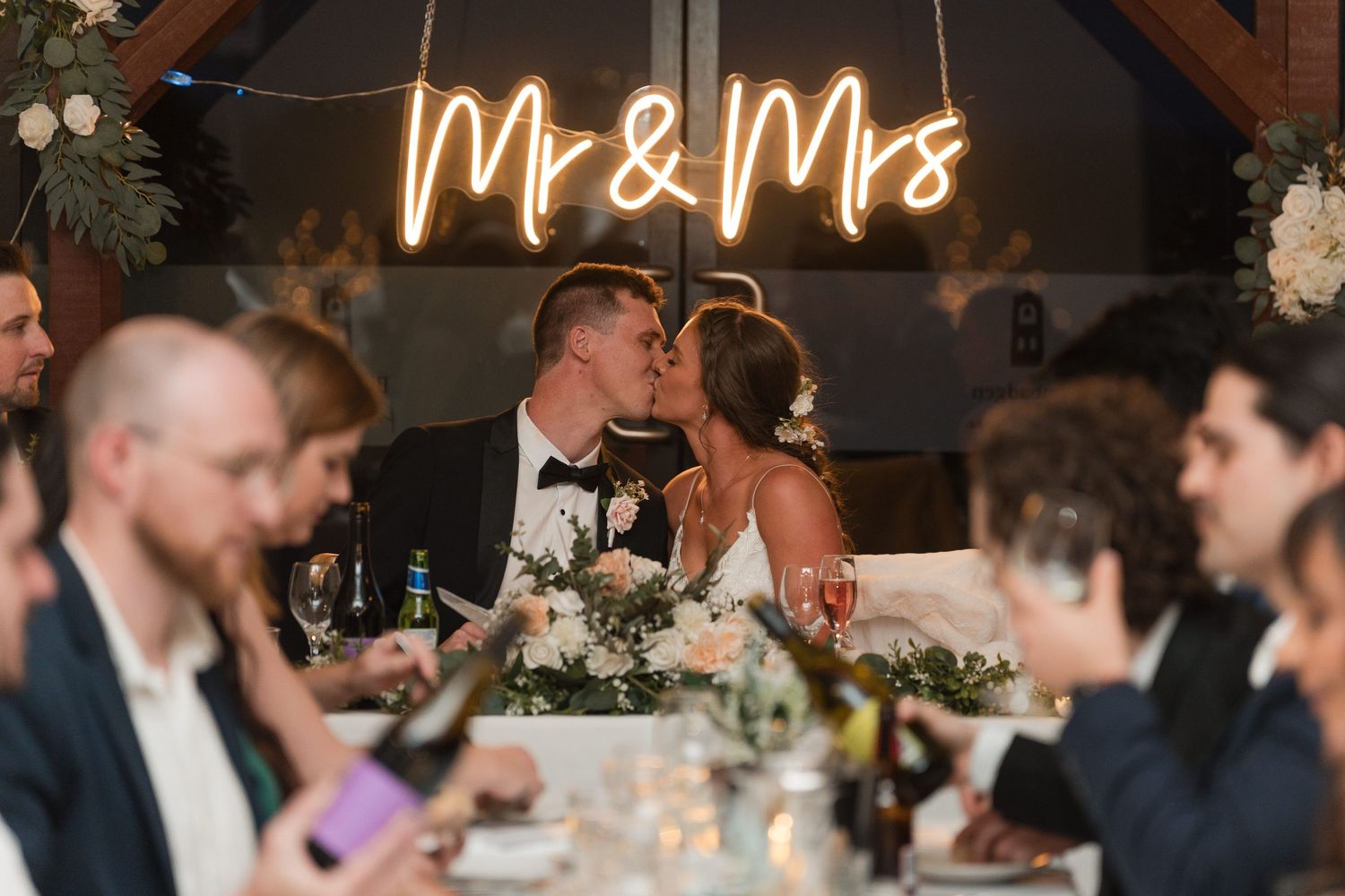 Bride and groom kissing at wedding reception under glowing Mr & Mrs neon sign surrounded by guests.