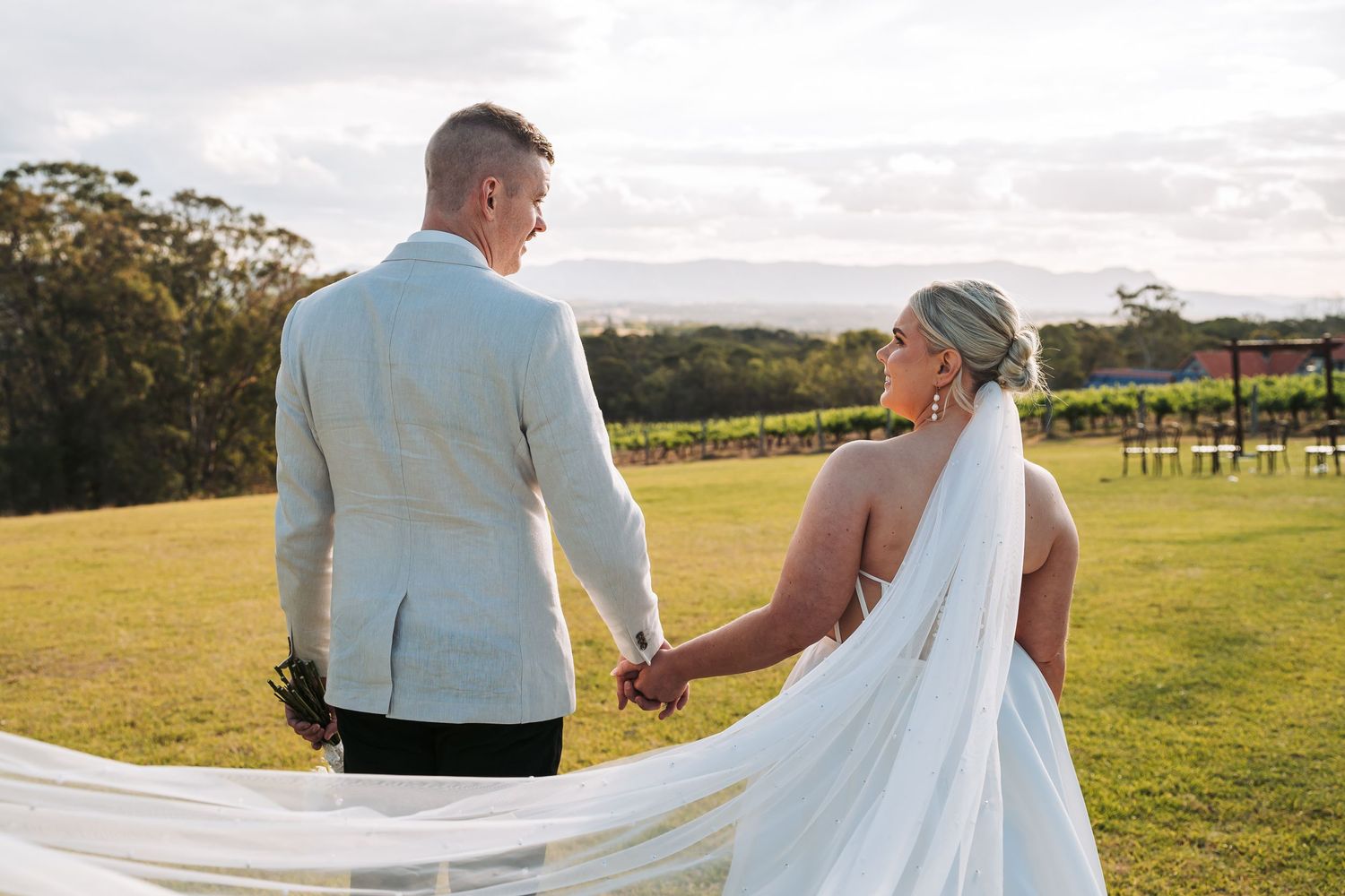 Newlyweds hold hands while walking through a scenic vineyard setting during golden hour.