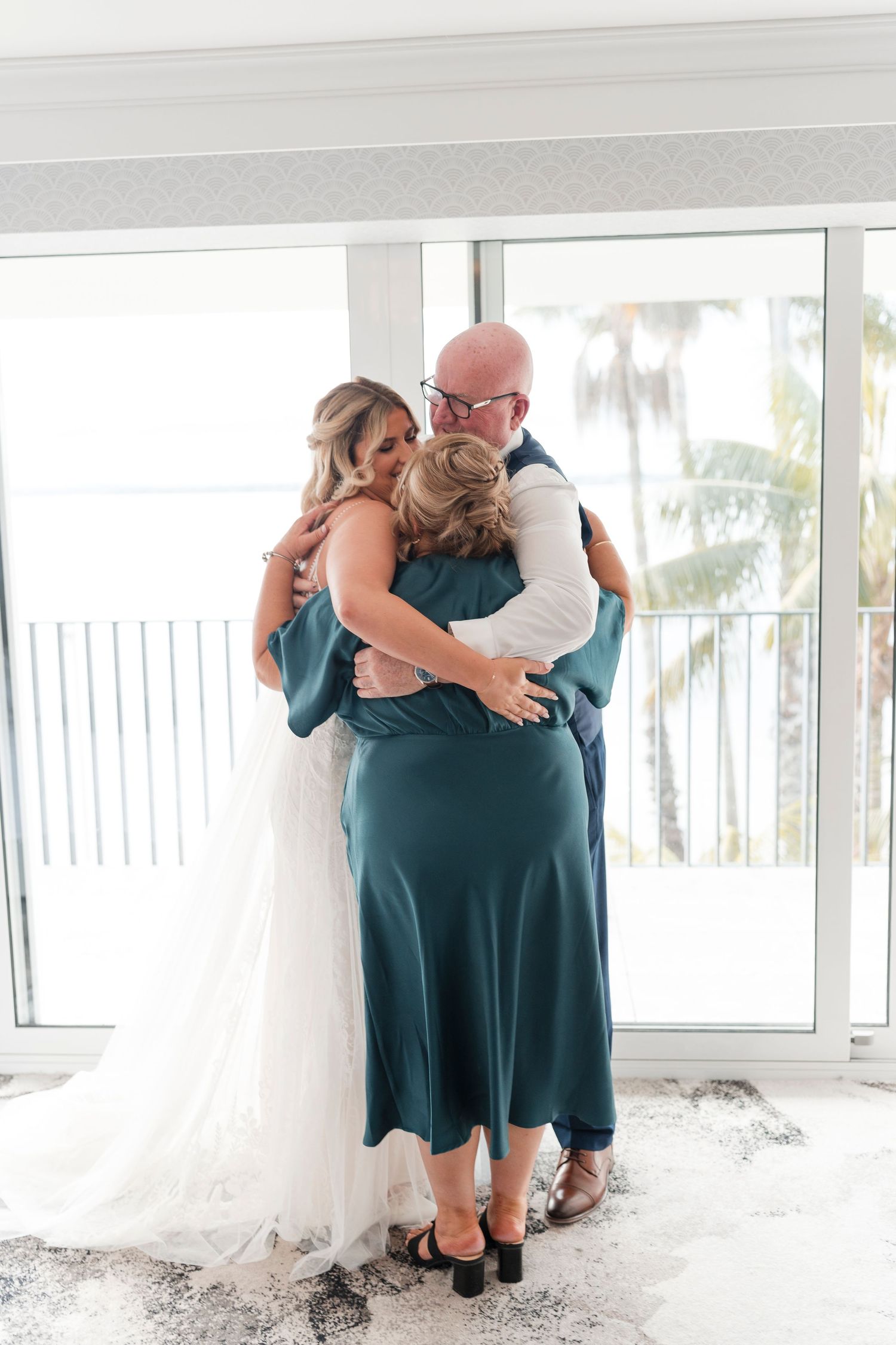 Emotional group hug between people in formal attire by a balcony window.