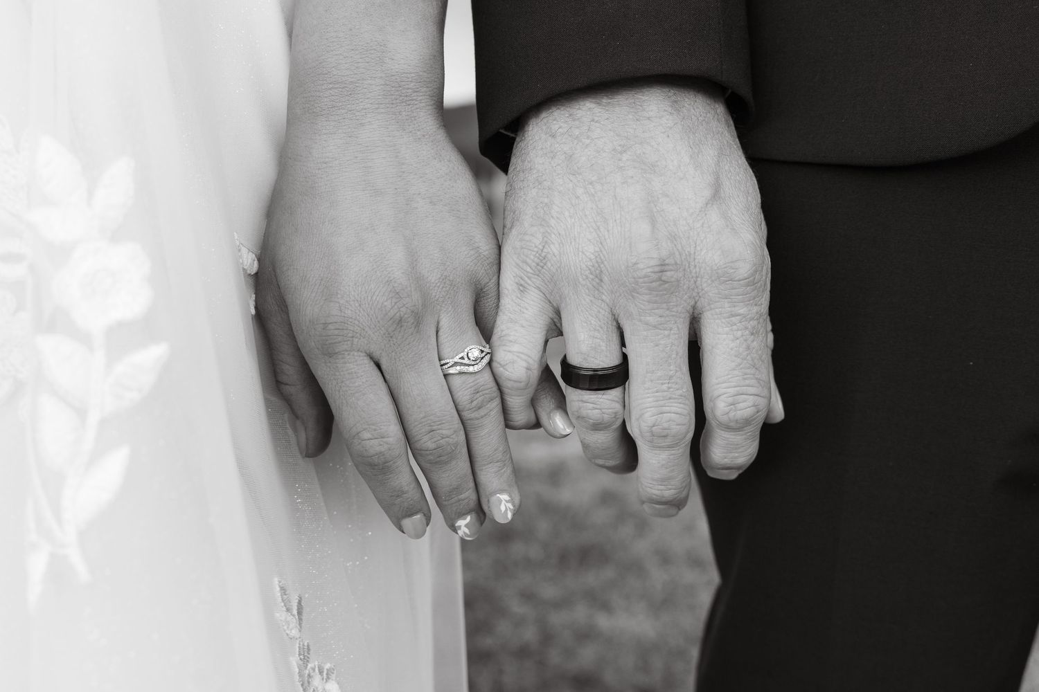 Black and white photo of bride and groom hands showing wedding rings, close-up detail shot.