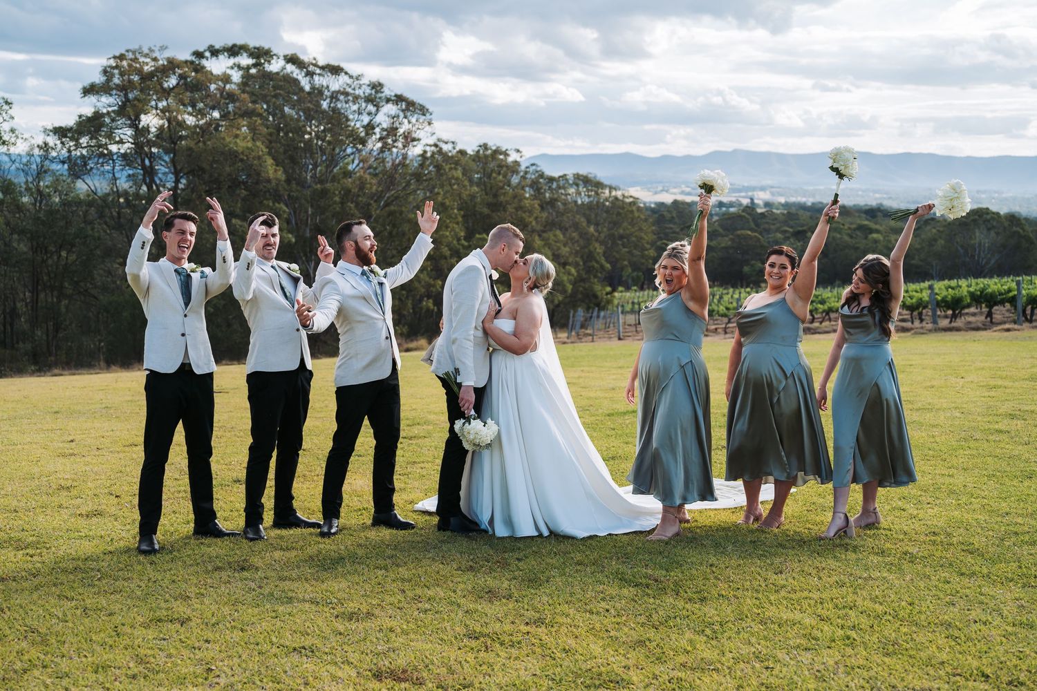 A wedding party poses joyfully on a grassy hilltop with mountains and cloudy skies in the background.