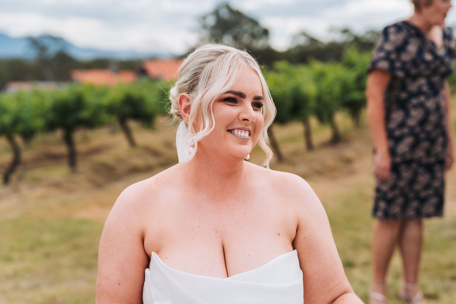 An outdoor portrait of a smiling person in a white dress standing in a vineyard setting.