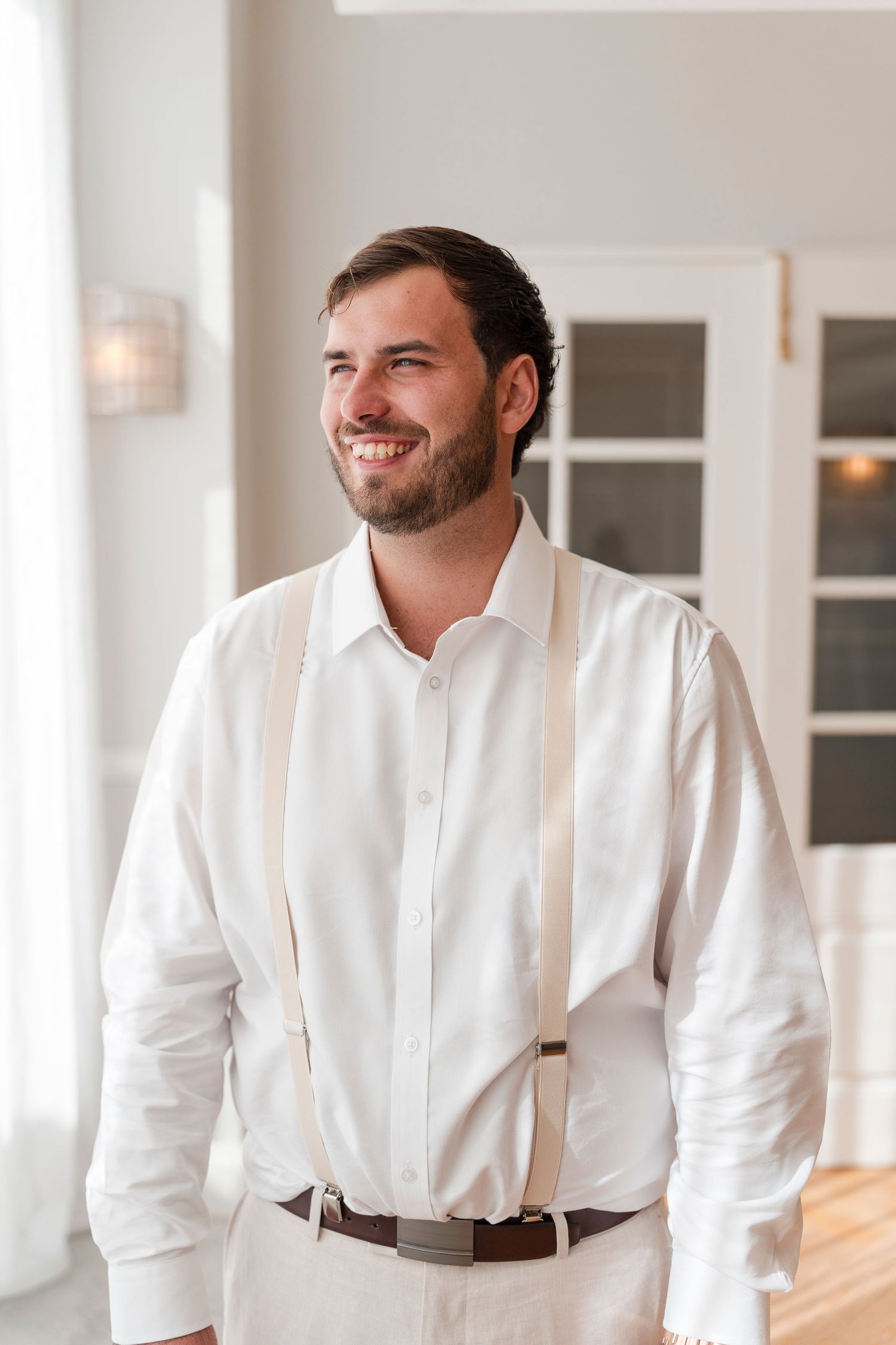 Person in white dress shirt and suspenders smiling in bright indoor setting.