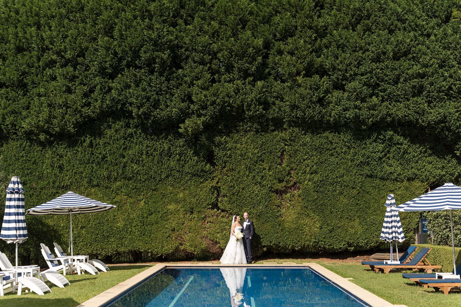 Bride and groom standing by a pool with striped umbrellas and lush green hedge backdrop at outdoor wedding venue.