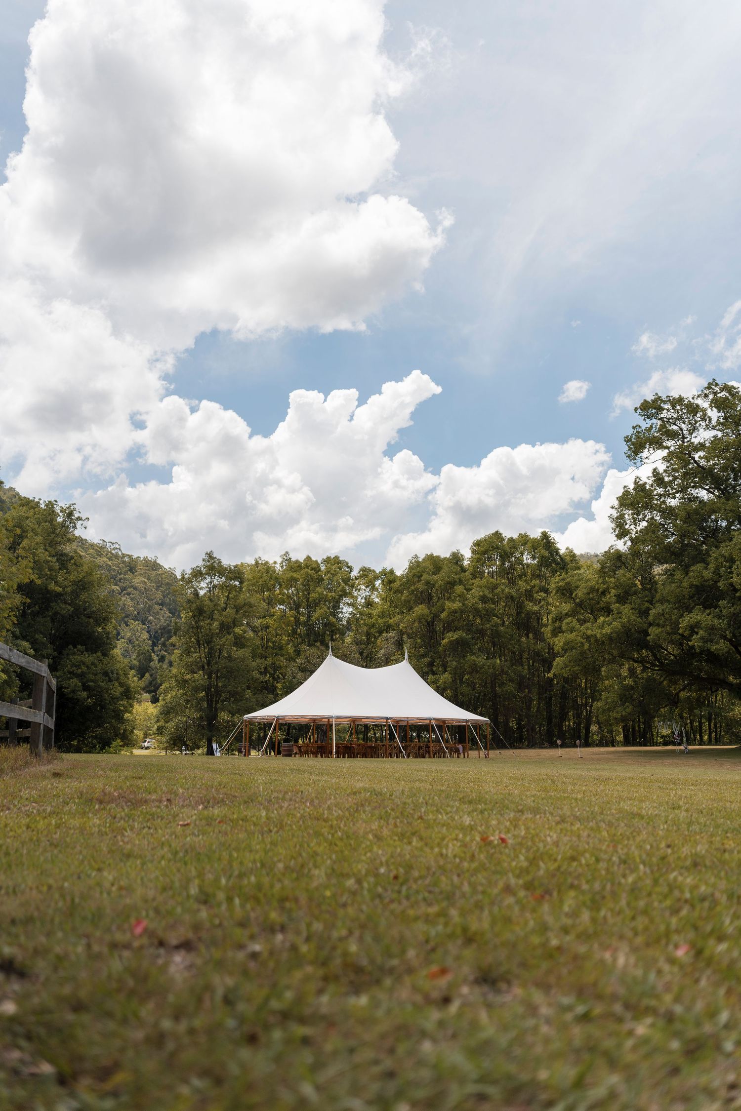 A white event tent stands in a grassy field surrounded by tall trees under a cloudy summer sky.